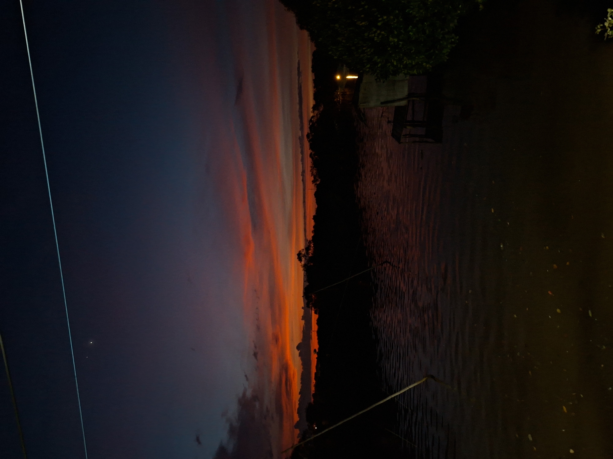 The image captures a serene twilight scene over a body of water, likely a river or coastal area in Lhokseumawe, Indonesia. The sky is a gradient of deep blues and purples, transitioning to vibrant oranges and reds near the horizon, indicative of a sunset. Several thin, wispy clouds are visible, catching the last rays of sunlight. A bright star or planet is visible in the upper left quadrant of the sky. A power line or thin wire stretches horizontally across the upper portion of the frame.
In the foreground, the water is dark and reflects the colors of the sky, with ripples suggesting a gentle current. Scattered leaves or debris float on the surface. Two thin, dark poles emerge from the water on the left side, suggesting some form of structure or marker.
The middle ground features a silhouetted line of trees and vegetation forming the distant shoreline. On the right side of the water, a small, dilapidated wooden structure with a table and chairs is partially submerged or very close to the water's edge. A bright, artificial light source illuminates the water near this structure, possibly from a distant house or floating object. Dense green foliage is visible on the far right, obscuring part of the scene. The overall atmosphere is peaceful and tranquil, with the vibrant sky contrasting with the dark foreground and silhouetted elements.