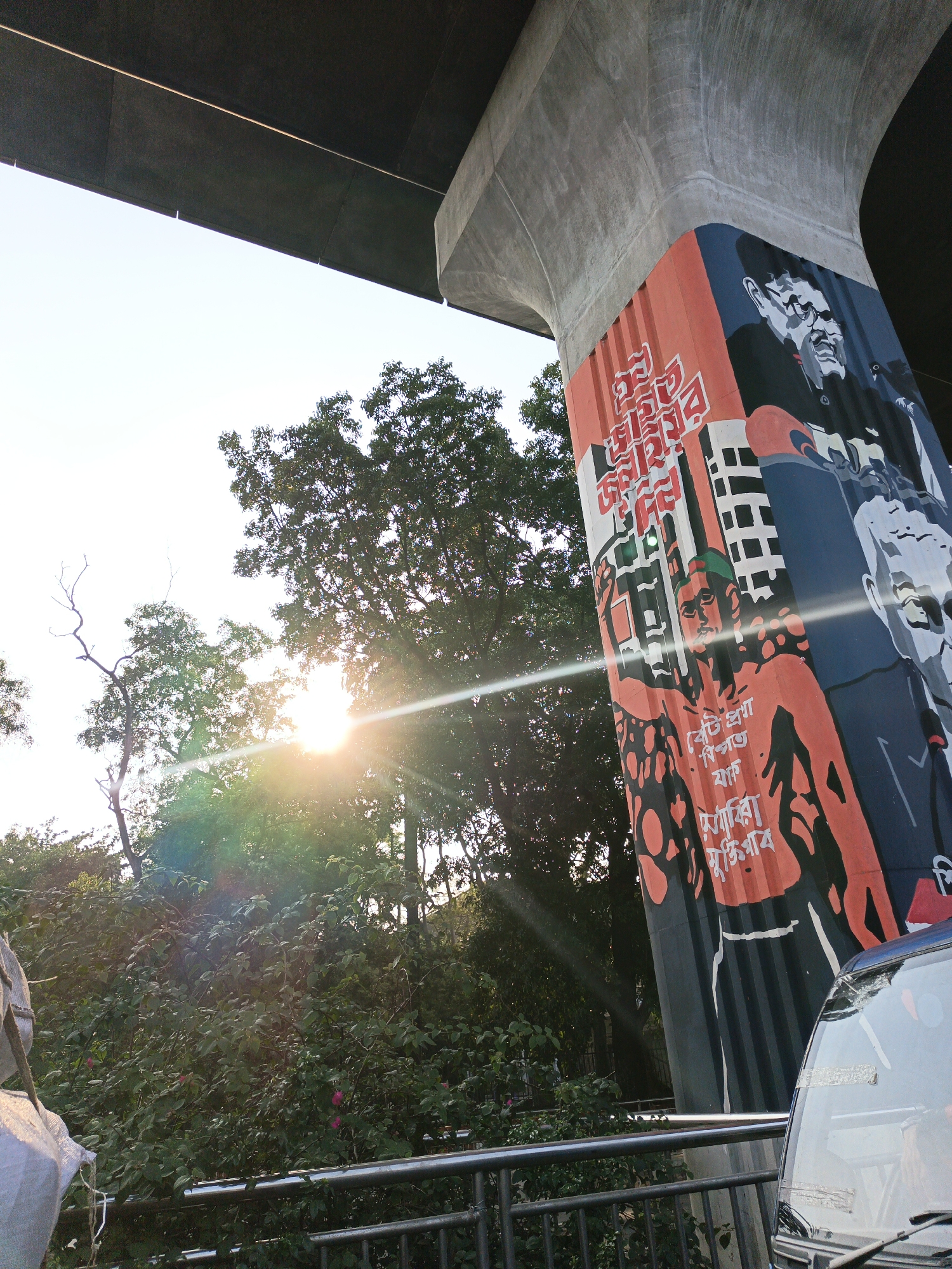 Captured from a low-angle perspective, the image showcases a scene from Unknown, Unknown. Sunlight streams through verdant trees, one notably sparse, contrasting with the artificial structures. A concrete pillar of an elevated roadway is adorned with a mural depicting portraits and calligraphic text in shades of red, black, white, and blue. A segment of the roadway above looms in shadow. The foreground includes a metallic railing and the partial view of a blue vehicle.