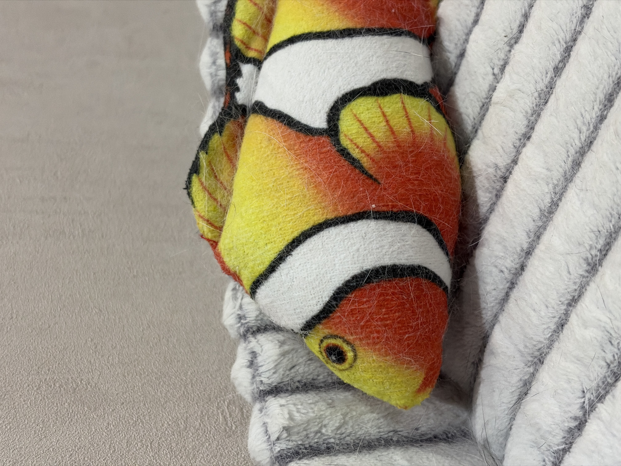 A close-up image shows a plush clownfish toy partially resting on a gray and white striped plush fabric. The clownfish is yellow and orange with white stripes outlined in black, and a black eye. The background consists of a light gray surface. The location in the city of Izhevsk, Russia is not discernible from the image.