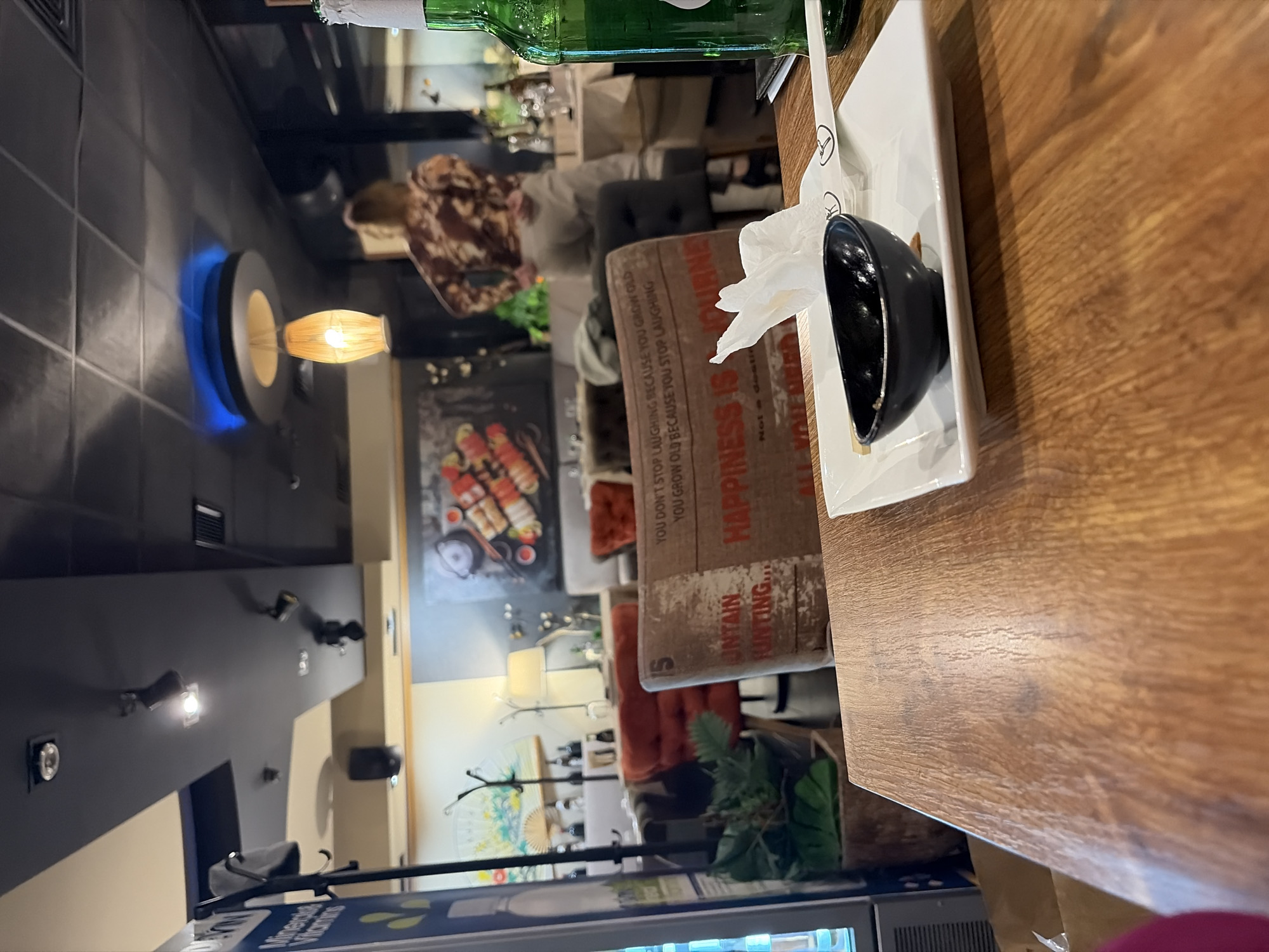 This image depicts the interior of a restaurant or cafe, identified by the prompt as being in Sofia, Bulgaria. The exact street address or name of the establishment is not discernible.
The perspective is from a seated position at a wooden table in the foreground. On this table, a white square plate holds a small black dipping bowl with some residue, a white paper chopstick wrapper with a circular logo containing the letter 'K' and a stick figure, and a crumpled white paper napkin. A green glass bottle is partially visible on the right edge of the table. A blurry pink object is in the lower-left corner.
In the midground, a chair with its back to the viewer is prominent. It is upholstered in a light brown fabric printed with text, including phrases like "YOU DON'T STOP LAUGHING BECAUSE YOU GROW OLD YOU GROW OLD BECAUSE YOU STOP LAUGHING" and "HAPPINESS IS A JOURNEY Not a desti...". Behind this chair, various seating arrangements are visible, including red upholstered armchairs and dark grey upholstered chairs, suggesting multiple dining areas. Tables with white tablecloths are set in the background.
A single person is observed in the background, standing with their back partially to the viewer, near a table. This individual wears a brown and white patterned long-sleeved top and light-colored trousers, with light brown or blonde hair pulled back.
The environment features a mix of wall colors, including light beige and darker grey/blue sections. Decorative elements include a large painting of sushi rolls on a dark wall, a large decorative fan with a landscape design, and green plants in pots. The ceiling is multi-level, with sections of dark and light panels, featuring recessed spotlights, track lighting, and a distinctive circular ceiling fixture emitting a blue glow, from which a woven pendant lamp hangs. A metal coat rack with a grey item hanging on it is visible on the left, next to a partial view of a commercial cooler or refrigerator with a blue-and-yellow label.