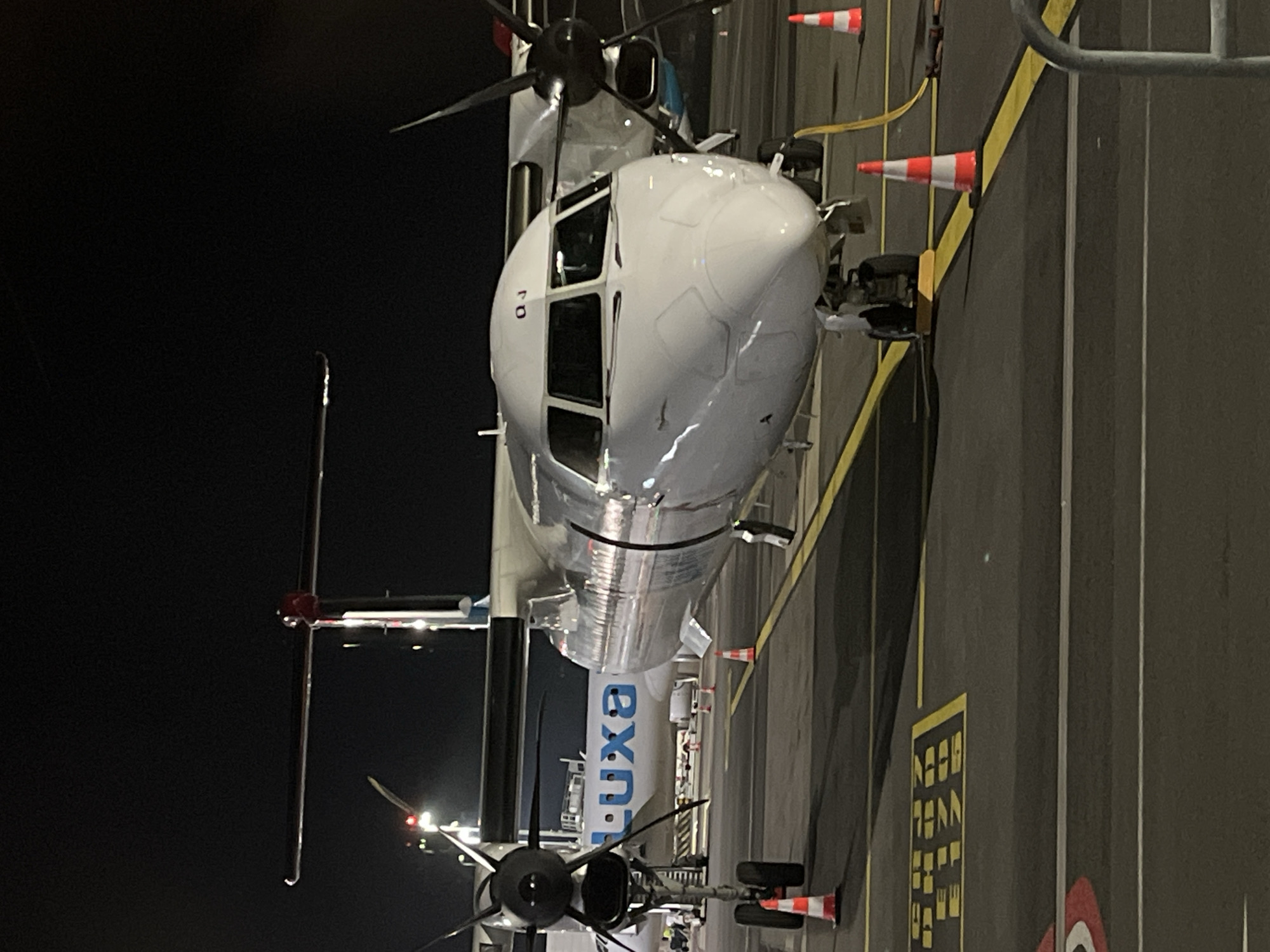 The image displays the front and mid-section of a white turboprop aircraft, likely an airliner, parked on an airport tarmac at night in Niederanven, Luxembourg. The aircraft's rounded nose and cockpit windows are prominent, reflecting artificial light. Two turboprop engines, each with visible propellers, are positioned on the wings. The main fuselage is white, transitioning to a metallic silver mid-section, with the blue letters "LUXA" clearly visible on its side, identifying it as "Luxair." A small identifier, possibly "QJ" or "GJ," is visible near the cockpit window.
The aircraft is stationary, indicated by its extended landing gear. The scene is an airport ground area, characterized by dark asphalt or concrete, bright yellow painted lines marking taxiways or parking stands, and several orange and white traffic cones placed around the aircraft. The very dark background confirms it is nighttime, with the aircraft and surrounding ground features illuminated by artificial lights. No people are visible, suggesting the aircraft is either parked overnight, undergoing maintenance, or awaiting its next operation.