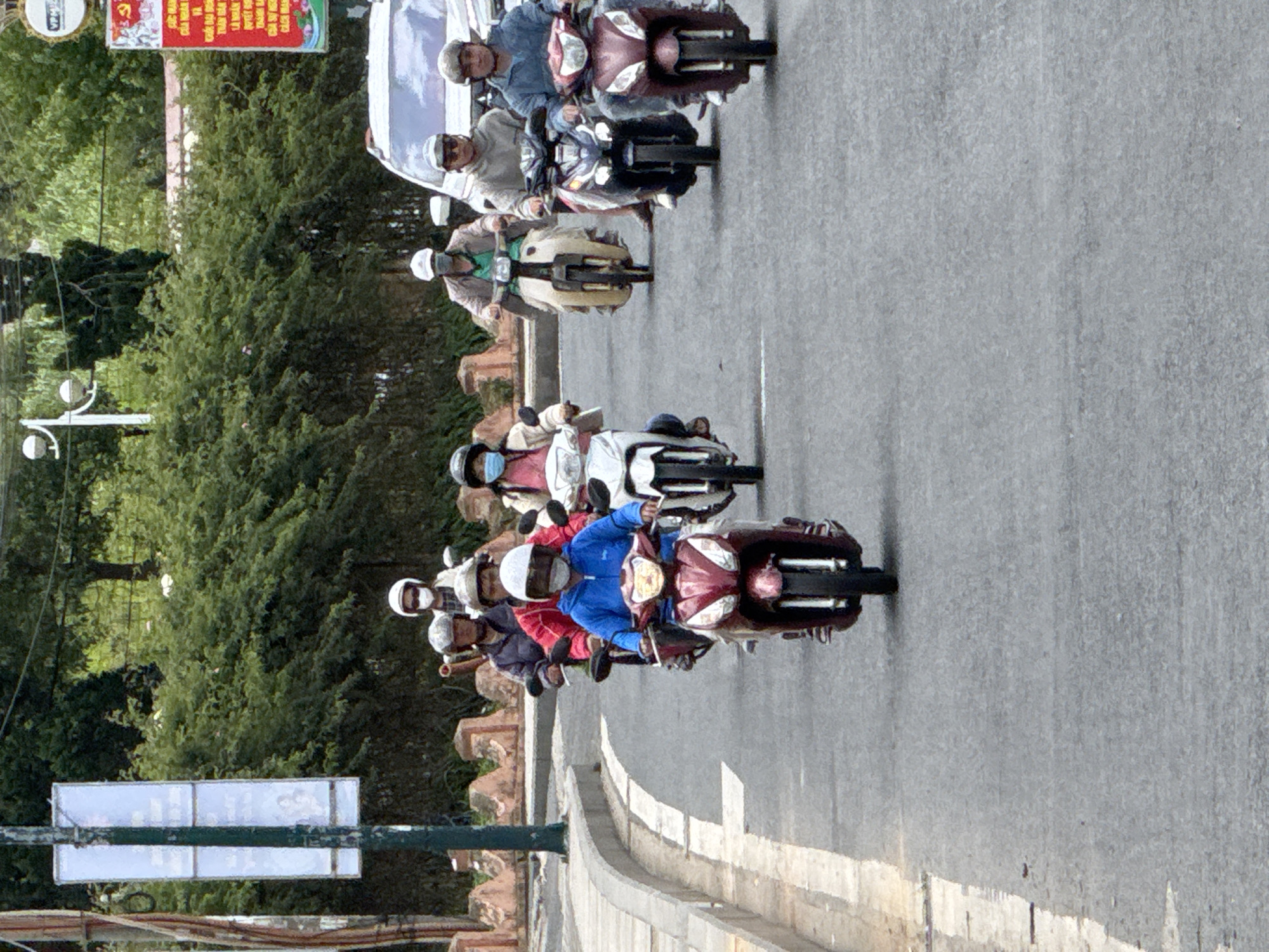 A group of people are riding motorbikes down a paved road in Da Lat, Vietnam. The road curves to the left and is lined with trees and some decorative stone walls. The people are wearing helmets and masks, and some are wearing jackets. The weather appears to be overcast. A white van is visible in the background. There are also signs and utility poles visible along the side of the road. The scene depicts a typical street scene in a Vietnamese city.