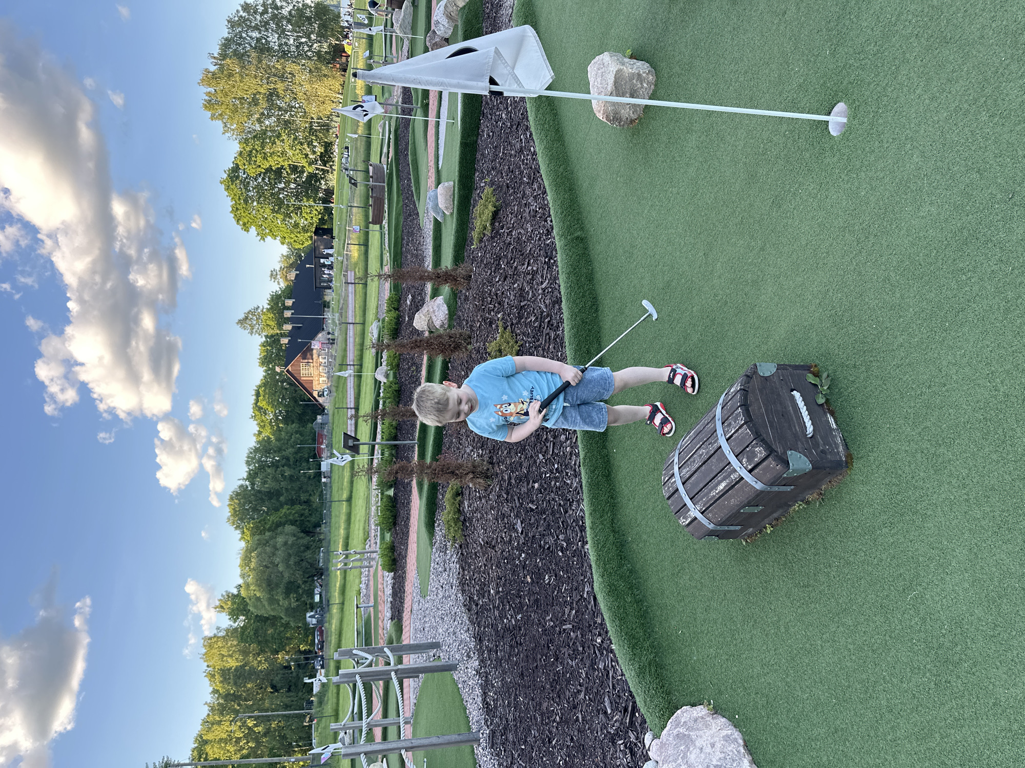 A young boy, appearing to be around 5 years old, is playing miniature golf. He is wearing a blue t-shirt with a character graphic, denim shorts, and red and black sandals. He is holding a putter and looking down at the golf ball on a green artificial turf putting surface. In front of him is a decorative wooden treasure chest, and to his right is a miniature golf flag. The background shows a miniature golf course with various obstacles, landscaping, trees, and a building. The sky is blue with white clouds, suggesting it is daytime.