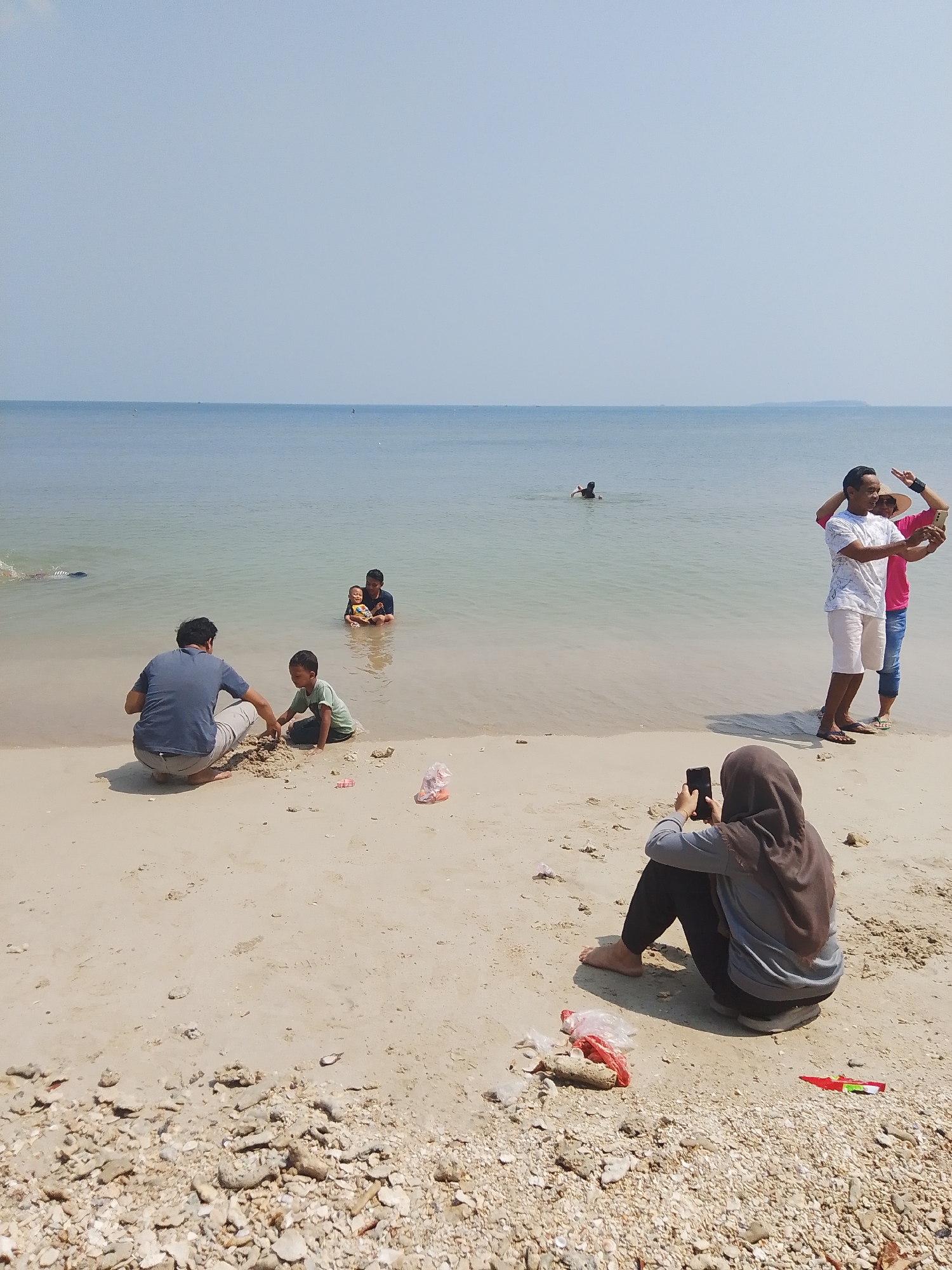 The image depicts a beach scene in Pejagan, Indonesia, with several people engaged in various activities. In the foreground, a person wearing a hijab is seated on the sand, holding a smartphone and appearing to take a photo. To the left, a man in a blue shirt and beige pants is crouching down, interacting with a young boy in a green t-shirt who is playing with sand. In the shallow water to the left, a person is swimming. Further out in the water, a child is being held by an adult sitting down, with another adult visible behind them. On the right side of the image, two men are standing in the water and taking a selfie with a smartphone. The beach is sandy, with scattered shells and some plastic debris visible on the ground. The ocean stretches out to the horizon under a clear, pale blue sky.