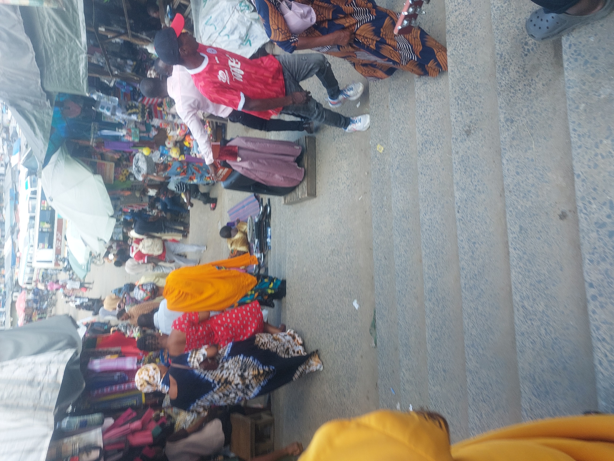 This media file presents a bustling outdoor market scene, likely in Dar es-Salaam, Tanzania, viewed from a slightly elevated vantage point. The atmosphere is vibrant and crowded, characteristic of a busy street market.
Numerous people populate the scene, engaged in various activities. In the foreground, towards the right, a man wearing a red football jersey with the partially visible text "MBE" and a logo, dark pants, and a red baseball cap stands. Beside him, another person is visible in a light pink long-sleeved shirt. Further right, an individual is seated on concrete steps, holding a guitar or similar stringed instrument, and adorned in a patterned garment. A foot clad in a grey Croc shoe is also seen on the steps.
The market itself is filled with vendors and shoppers interacting among makeshift stalls. These stalls are often covered by light-colored tarpaulins (grey and white) and display a diverse range of merchandise. Visible goods include colorful clothing such as red and orange tops, as well as patterned fabrics. Other items like a white umbrella, a small fan, and a water bottle can be observed. People are seen walking, standing, sitting, and tending to their wares.
The ground is a textured concrete or paved surface, with distinct steps forming the right edge of the frame. The lighting suggests it is daytime, possibly under an overcast sky or diffused sunlight, as strong shadows are absent. The overall scene conveys an active and lively urban market environment focused on commerce and daily life.
