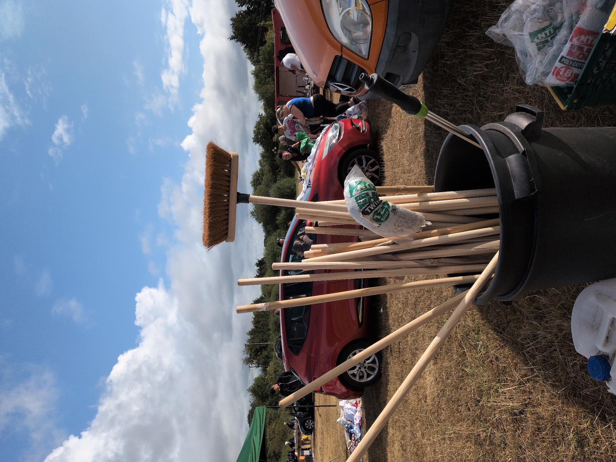 A wide-angle outdoor shot depicts a sunny day at what appears to be a car boot sale in South Staffordshire, United Kingdom. The foreground is dominated by a large black bucket filled with numerous wooden broom handles and a broom head. A white plastic container with a blue lid sits in the lower left. Behind the bucket, a red hatchback car is parked on dry grass, its doors open, with several people milling around it. To the right, the front of an orange van is visible, along with a green plastic crate containing bagged items. In the background, a green gazebo is set up, and more people and cars are scattered across the field, suggesting a lively event. The sky is a clear blue with scattered white clouds.