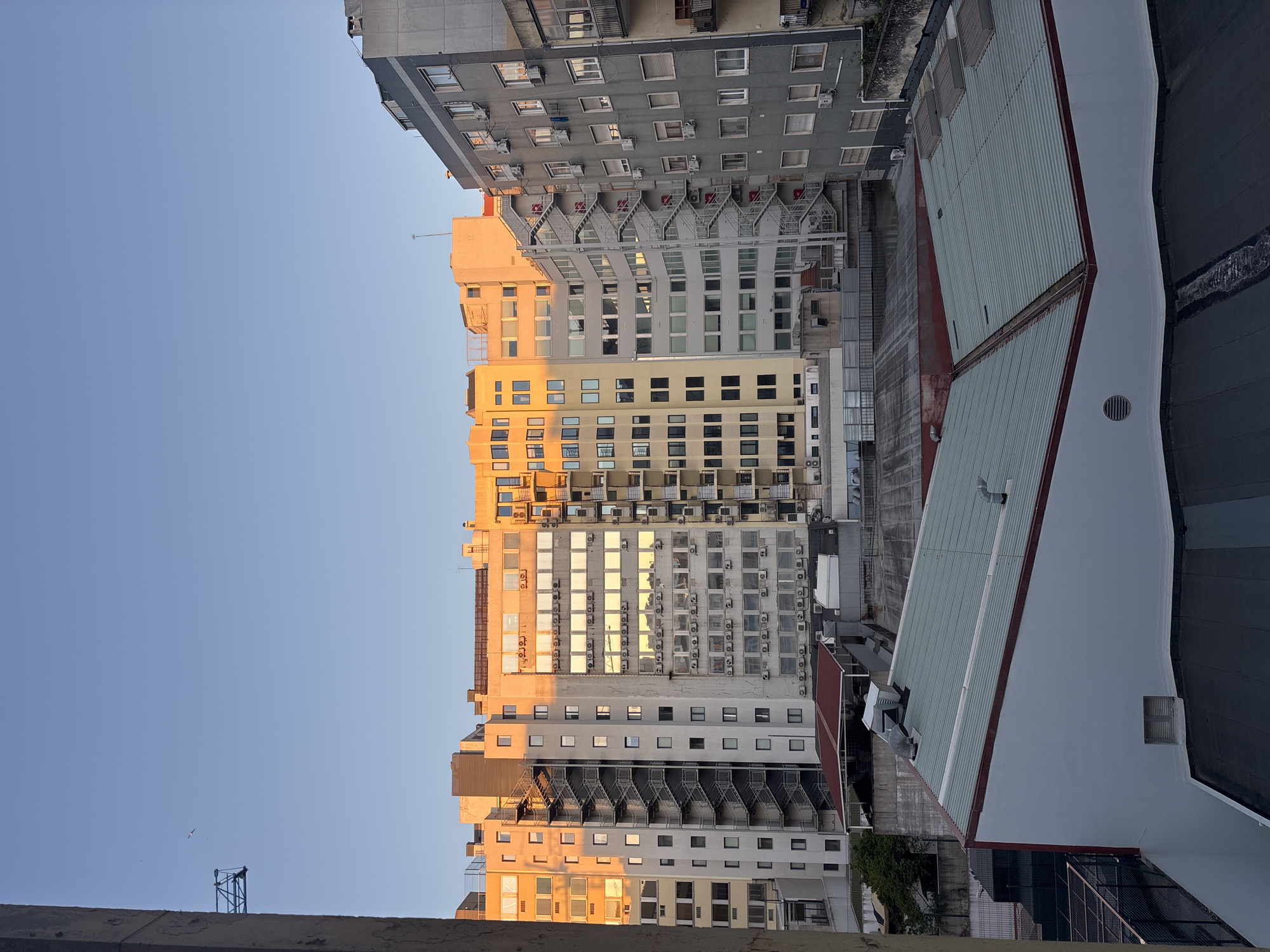 The image depicts a view of urban buildings in Lisbon, Portugal. The buildings are predominantly light gray and yellow with numerous windows, some reflecting sunlight. Several buildings have exterior air conditioning units. Fire escapes are visible on some buildings. A section of a large, white and grey-green roof with a red edge occupies the right side of the image. The sky is a clear blue. A small crane is visible in the lower left.