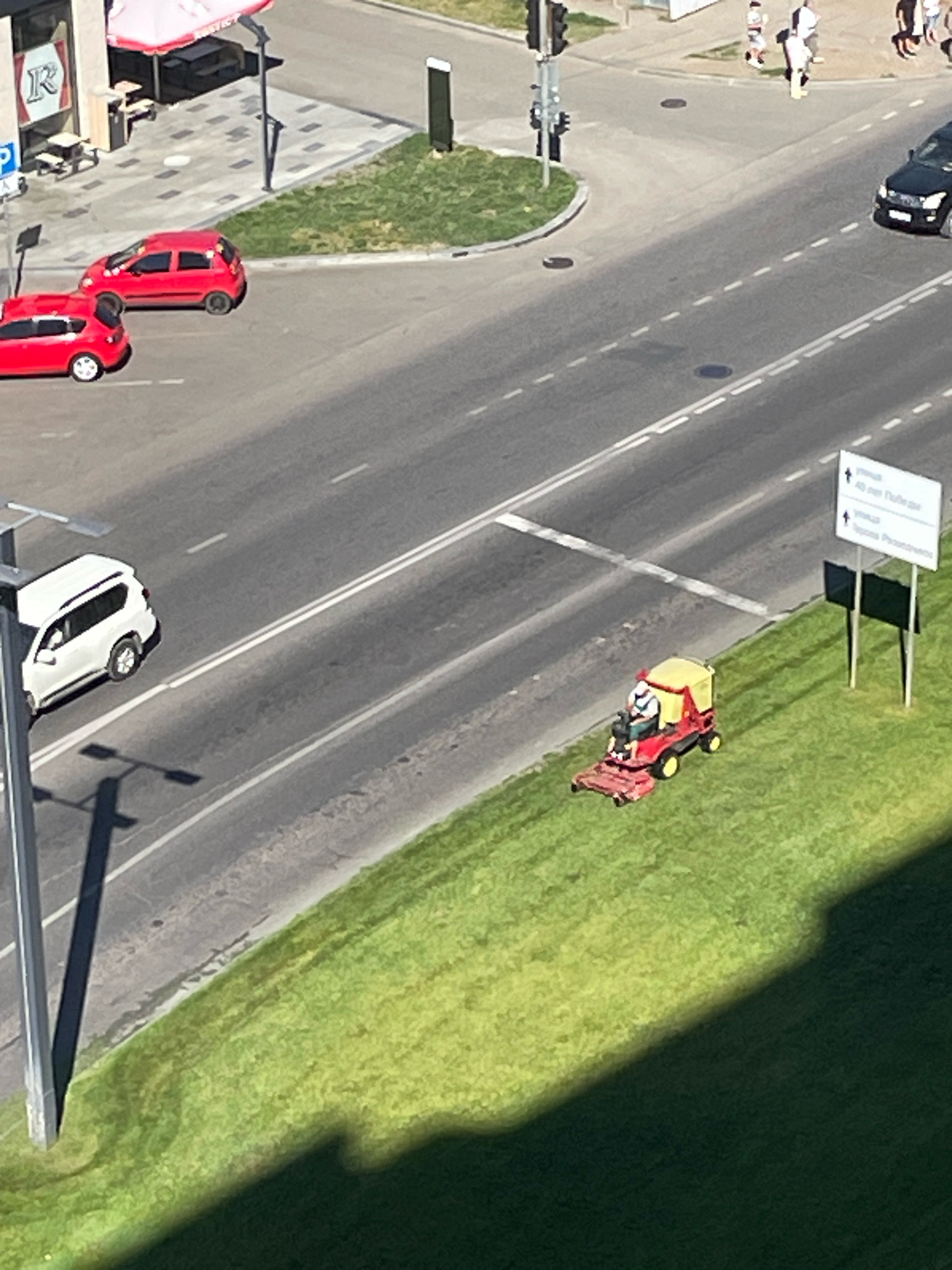 An aerial, high-angle, daylight, medium shot shows a street scene in Krasnodar, Russia. A person is operating a red and yellow ride-on lawnmower on a patch of green grass next to a road. The person is wearing a white shirt and blue pants. The road has double white lines painted down its center and is currently devoid of traffic, except for a white SUV driving away in the distance on the left, and a black SUV driving away on the right. In the background, there is a building with red awnings and tables outside, and a traffic light pole. To the right of the lawnmower, a white directional sign with black lettering is visible.