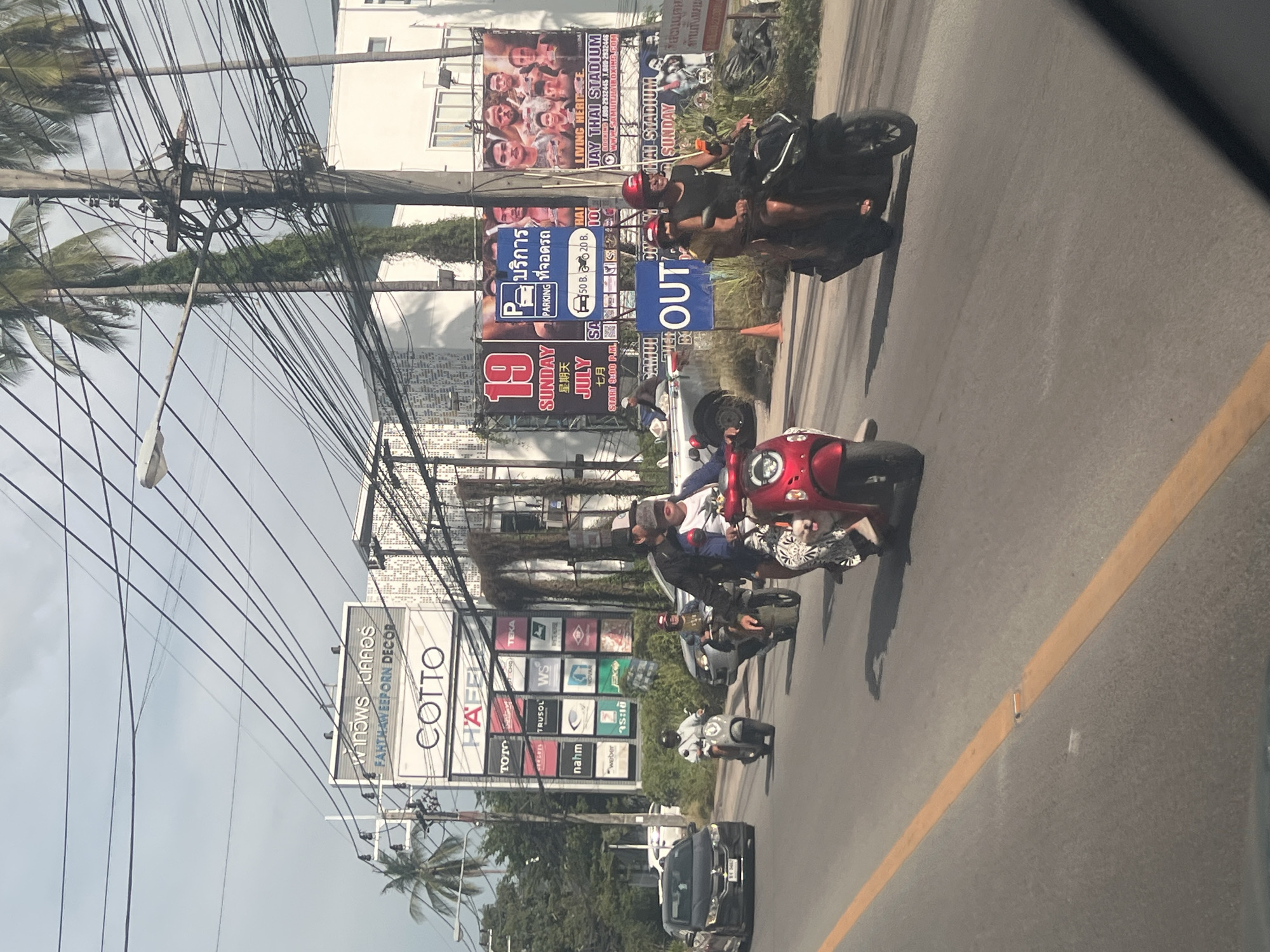The image depicts a street scene in the Ko Samui District of Thailand. Several motorcycles are present on a paved road. Two motorcycles are in the foreground, one red and one black, each carrying two people. The people on the red motorcycle are a woman in the front wearing a patterned dress and a man in the back wearing a dark shirt and a red helmet. The people on the black motorcycle are a man in the front wearing a black shirt and a black helmet, and a woman in the back wearing a patterned dress. A gray scooter with a single rider wearing a white shirt and a dark helmet is visible further down the road. A dark-colored car is in the left lane.
In the background, there are buildings with signage. One large billboard displays "19 JULY SUNDAY" in English and Chinese, along with images of boxers. Another billboard features "LIVING HERITAGE SUAY THAI STADIUM SUNDAY". Other signs include "FAHTHAWEEPORN DECOR", "COTTO", and various brand logos like "TOTO" and "WEBER". Palm trees line the street, and utility poles with a dense network of wires are prominent. The sky is overcast. The road is marked with a yellow line. The overall environment suggests a commercial area on a street in Ko Samui.