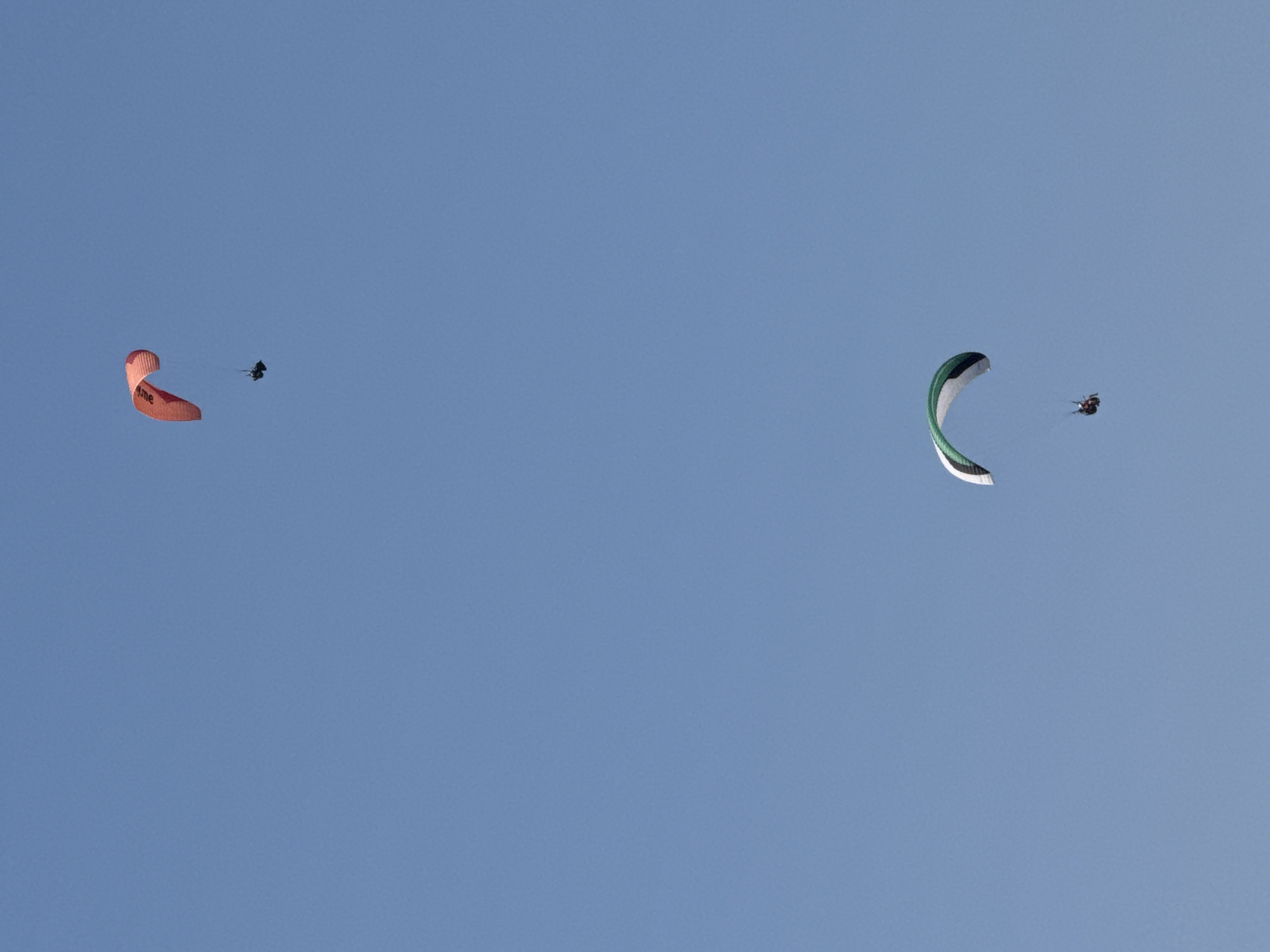 Two paragliders are visible in a clear blue sky. The first paraglider, positioned in the upper portion of the frame, has an orange wing and is descending. Below and to the right, a second paraglider with a white and green wing is also descending. Both paragliders have a single person suspended beneath each wing in a harness. The image is taken in the city of Boreti, Montenegro, and depicts an outdoor recreational activity.