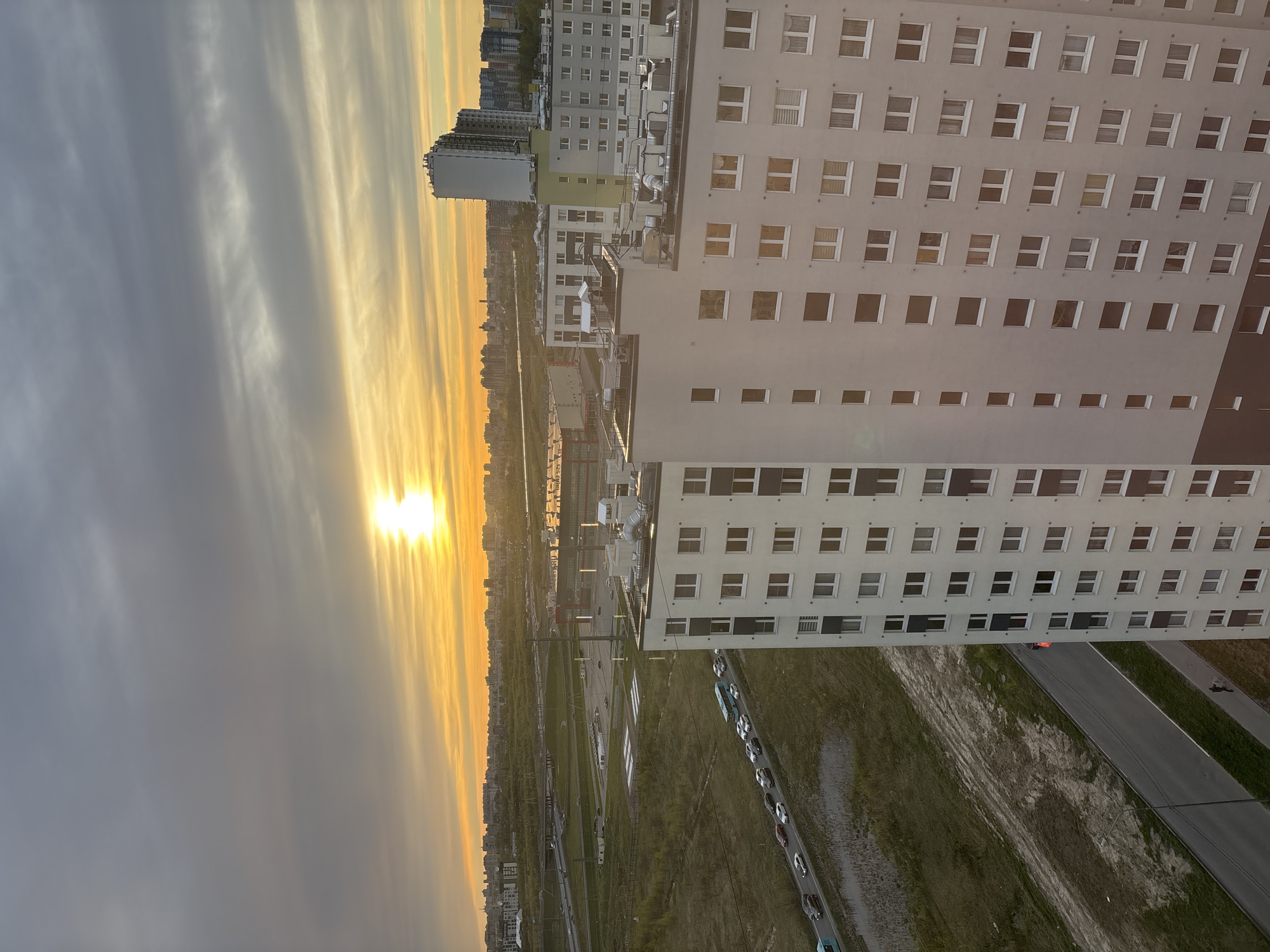 The image shows a high-angle view of Vsevolozhsk, Russia, at sunrise or sunset. The sky is filled with clouds tinged with orange and yellow. A tall, white apartment building dominates the right side of the frame. Numerous windows line its facade. To the left, other buildings are visible in the distance. Below the apartment building is a road with a line of parked cars, and a grassy area that appears to be a railway track. The sun is low on the horizon, casting a bright light. A small number of people can be seen walking on the ground.