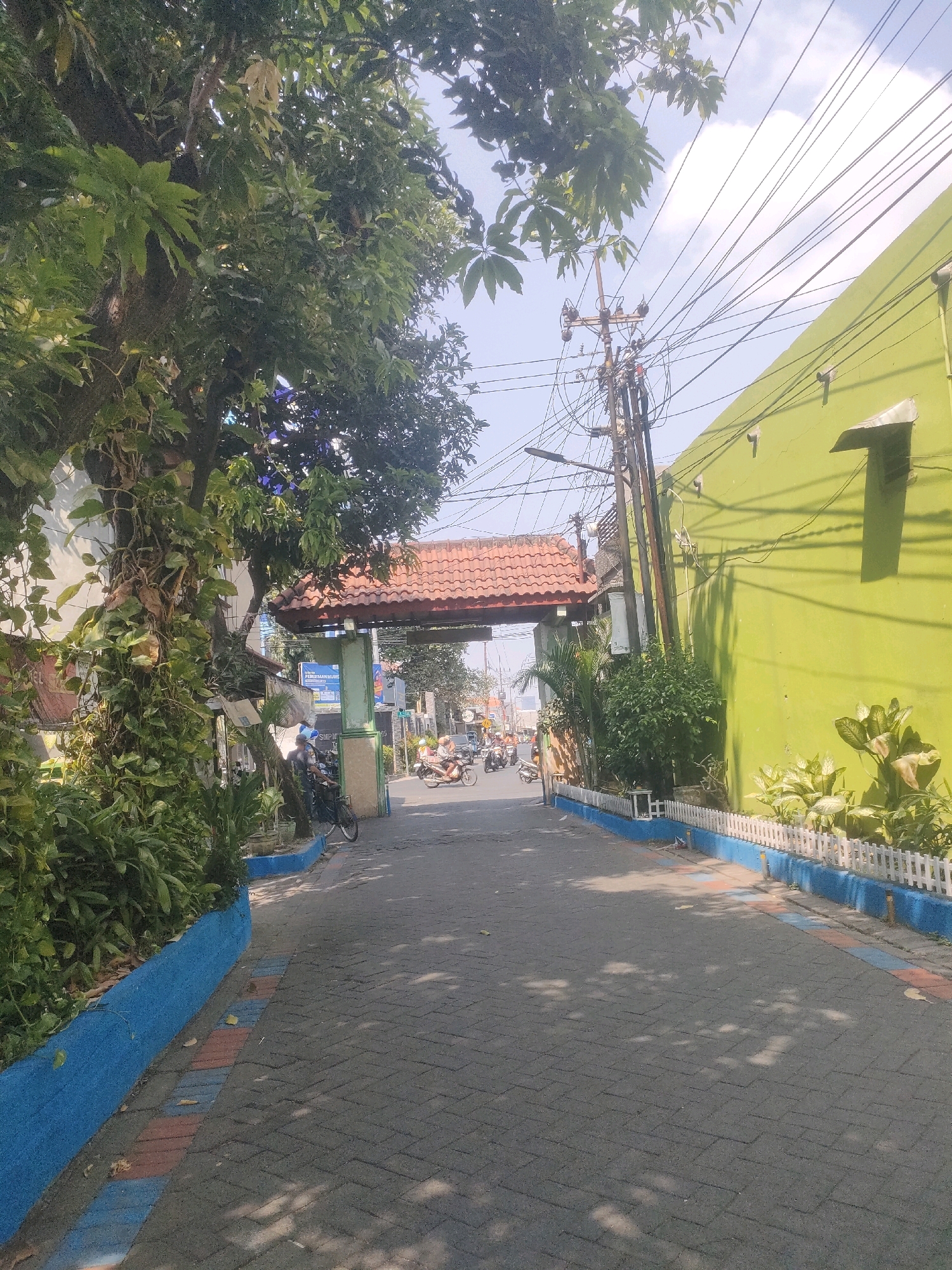 The image depicts a sun-drenched street in Surabaya, Indonesia, likely in a commercial or residential area with significant greenery and infrastructure. The foreground is dominated by a paved walkway constructed from dark gray interlocking bricks, interspersed with patches of red and blue. To the left, a large tree with lush green foliage and vines growing up its trunk provides shade. A low, bright blue wall runs along the edge of the walkway, separating it from a planter filled with dense green bushes and smaller plants. Further down the street, an ornate archway with a red tiled roof spans the road, marking an entrance or transition point. Beyond the archway, the street continues, with several motorcycles and a bicycle visible. On the right side of the street, a vibrant lime green building stands tall, its surface marked by shadows from overhead power lines. A white picket fence, bordered by a blue strip, encloses a garden area in front of the green building, featuring various green plants and some with light-colored leaves. A utilitarian utility pole with numerous wires is prominent in the middle ground, suggesting an urban setting. The sky above is a clear blue with scattered white clouds. The overall impression is that of a bustling, everyday scene in a tropical city.