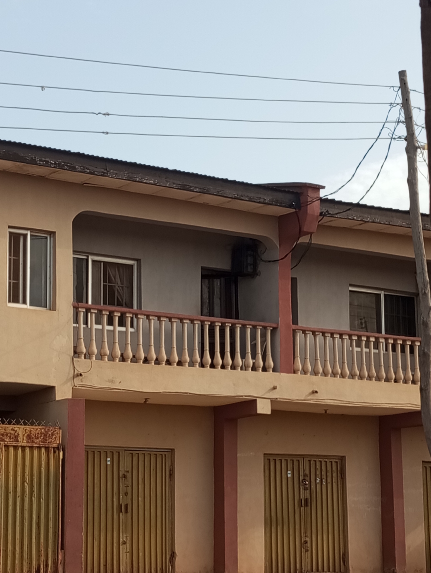 The image depicts a two-story building in Narayi, Nigeria. The building is painted in a combination of beige and reddish-brown colors. The top floor has a balcony with a balustrade. Windows are visible on both floors. The ground floor has metal security doors or gates. Overhead power lines are visible against the sky. A utility pole stands to the right of the building. An air conditioning unit is mounted on the wall of the upper floor. The lighting suggests daytime, likely under diffused sunlight.