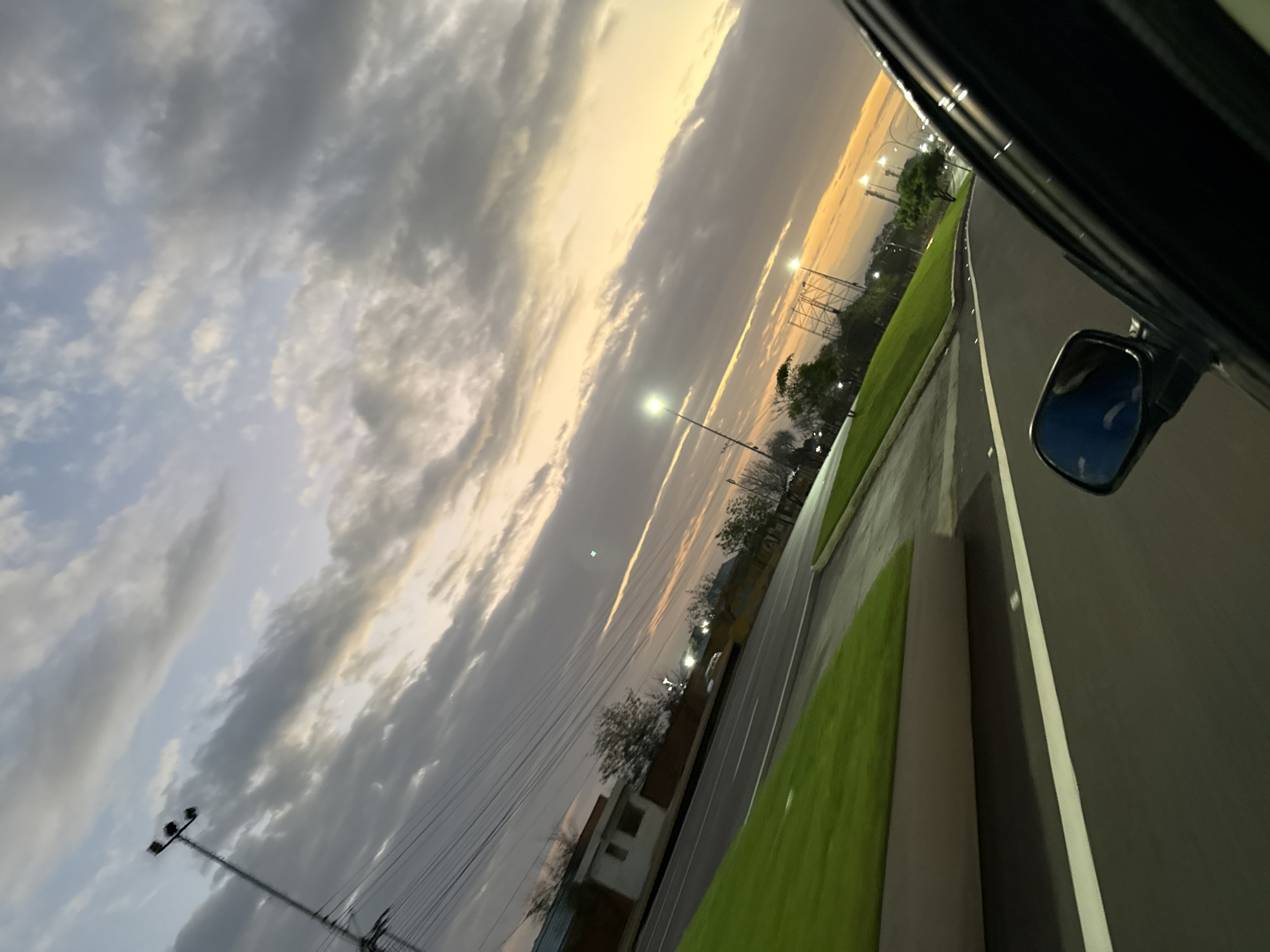 A slightly tilted shot taken from inside a car, likely the passenger seat, shows a road lined with green grass and trees heading toward a sunset in Unknown, Unknown. Street lights are visible along the road. A building is visible behind the trees. The side mirror of the car reflects a blue sky. The sky features a mix of clouds and yellow light from the setting sun. Power lines run alongside the road. The car door is visible in the foreground.