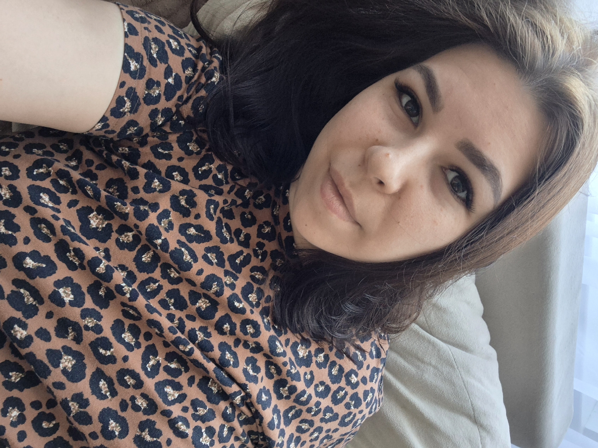 A woman with brown hair and brown eyes is lying on a beige surface. She is wearing a brown and black leopard print shirt. The location is Unknown, Unknown.