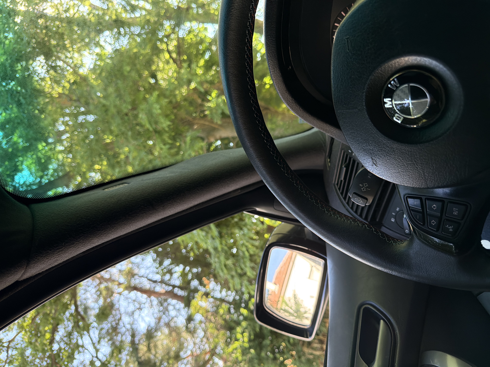 The image shows the interior of a car, looking out through the windshield and side window. The steering wheel is visible in the foreground, with the BMW logo prominently displayed on the center. To the left, a side mirror reflects a building with a red-tiled roof. Outside the car, lush green trees are seen, with sunlight filtering through the leaves, creating a bokeh effect. The scene suggests a sunny day, possibly in a suburban or rural setting, as indicated by the presence of trees and a house. There are no people visible in the image. The car appears to be parked or moving slowly. The location is identified as Dinkata, Bulgaria.