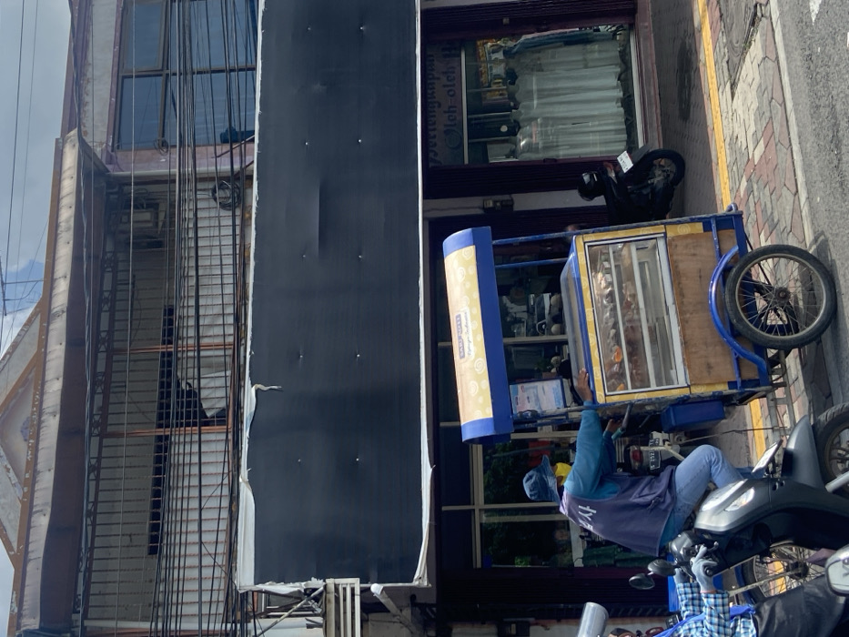 The image shows a street scene in Mojokerto, Indonesia, at the coordinates -7.467 latitude and 112.44 longitude. A man wearing a blue vest, a white shirt, and a blue cap is sitting on a motorcycle with a blue and yellow food cart attached. The cart is loaded with various food items, visible through a glass display. The man appears to be in the process of driving the cart, as he is looking ahead and his right hand is on the handlebar. Behind him, a storefront with a large black sign is visible. The sign reads "Setenganpra Oloh-oleh". In the storefront window, white garments can be seen. A motorcycle is parked in front of the store. The scene is set on a paved street with yellow painted lines. The sidewalk is made of grey bricks and a yellow painted curb borders the road. The weather is sunny.
