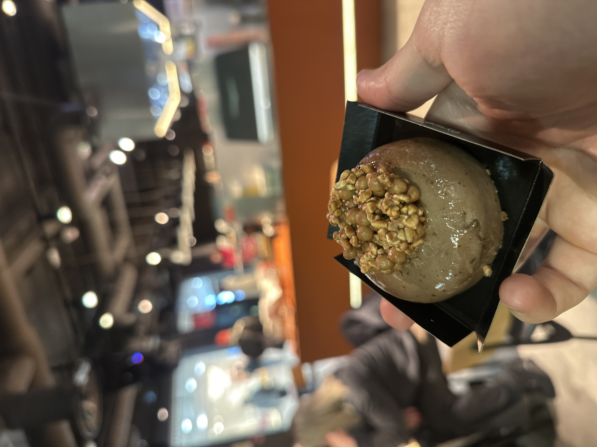 A hand holds a small, spherical dessert covered with chopped nuts in a square, black container. The dessert appears to be light brown or gray in color. The background shows a blurred interior scene with bright lights, suggestive of a public space like a shopping mall or restaurant. The image is taken at an oblique angle. No people are visible. It is not possible to definitively identify the location as Saint Petersburg, Russia from the image.