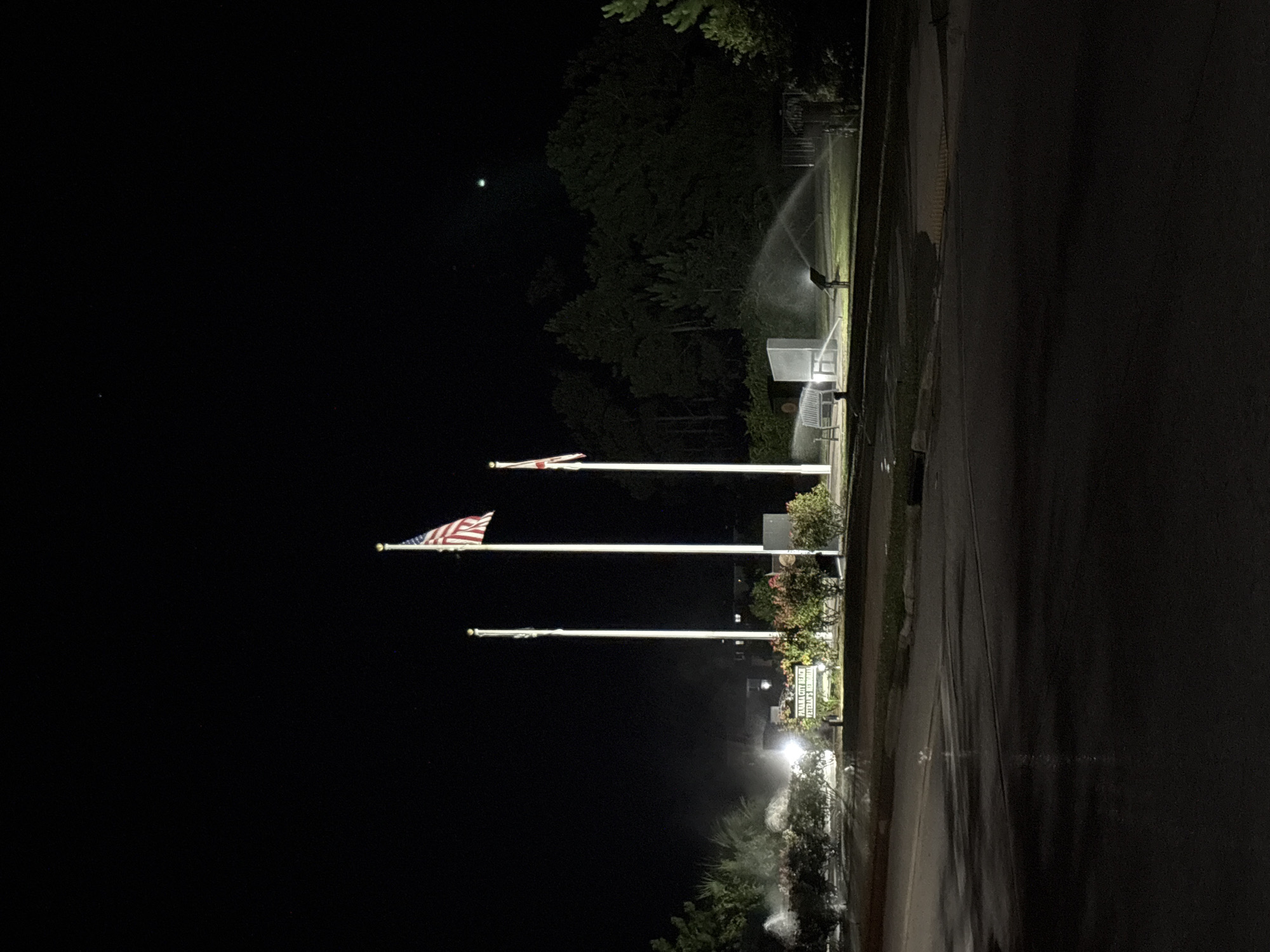 The image shows the Panama City Beach Veterans Memorial at night. Three flags are prominently displayed on tall poles. The leftmost flag is white with a red cross. The middle flag is the American flag. The rightmost flag is white with a red cross and a white field in the canton. The memorial features a white monument or plaque with text that is partially legible, reading "PANAMA CITY BEACH VETERANS MEMORIAL". In front of the memorial, a park bench is visible, and sprinklers are watering the surrounding green area. The scene is illuminated by artificial lights, creating a stark contrast with the dark night sky. The ground in the foreground appears wet, possibly from rain or the sprinklers. Trees and bushes form the background.