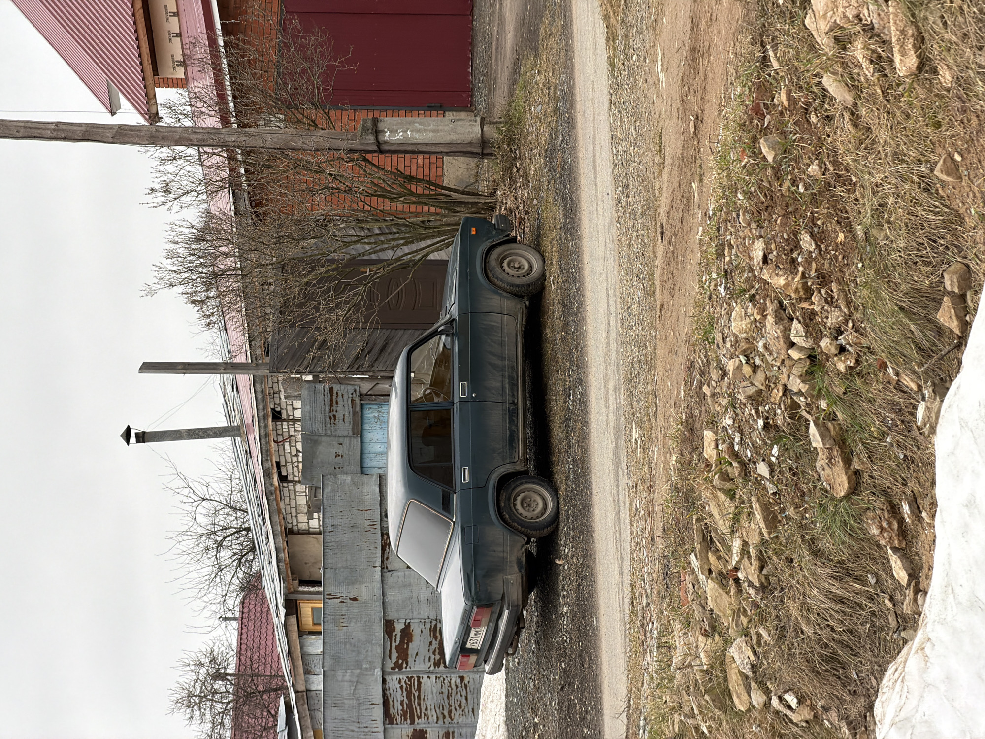 The image depicts a green Lada sedan parked on the side of a dirt road in what appears to be a semi-rural or urban fringe area. The car is parked close to a corrugated metal structure, possibly a garage or shed. A brick building is visible in the background, and there are trees with bare branches nearby. The ground is covered with dirt, rocks, and some vegetation. The license plate on the car is visible. The location is in the city of Chaikovsky, Russia.