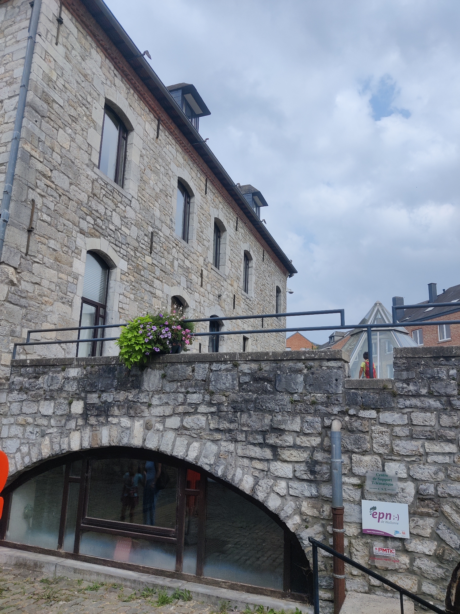 The image captures a view of a stone building in Marche-en-Famenne, Belgium, likely near the former prison which is now a cultural center. The building, constructed with rough-hewn stones, has several arched windows and a steeply pitched roof with dormer windows. A metal railing runs along a stone ledge in front of the building, adorned with a large pot of flowering plants in shades of pink, purple, and green. Below this ledge, an arched window or doorway with reflective glass shows distorted reflections of people inside. A metal drainpipe runs vertically down the stone wall to the right of the window. Attached to the wall are several signs: one reads "Centre de Support Télématique - C.S.T.", another features a logo with "epn :'-) de Wallonie", and a third sign, lower down, displays "PMTIC". The sky is overcast with visible clouds.
