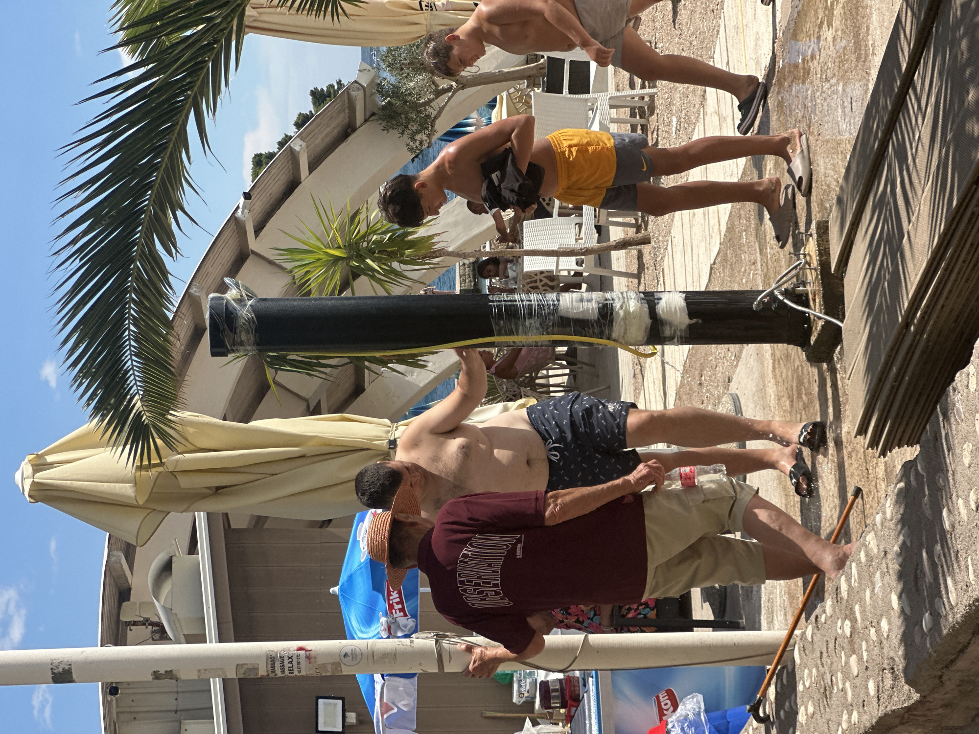 In Budva, Montenegro, on a sunny day, a group of people are gathered near a beachside establishment. An older man wearing a straw hat and a maroon shirt with the word "OBSERVATION" printed on the back is talking with a shirtless man in patterned swim trunks. They stand on a wet, cobbled surface. Nearby, two teenage boys, one in yellow shorts and the other in striped shorts, are also present. The boy in yellow shorts is holding dark clothing. Palm trees and umbrellas provide shade. In the background, there are tables and chairs set up, suggesting an outdoor seating area for a restaurant or bar. The overall atmosphere appears casual and relaxed, typical of a summer day at a coastal resort.