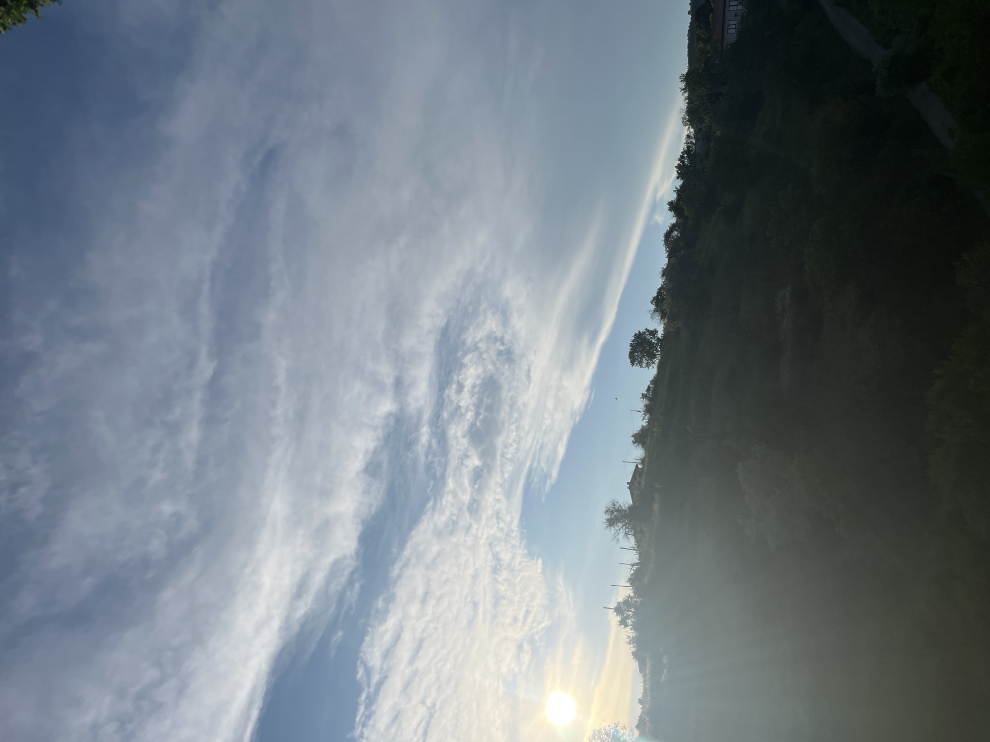The image captures a sunlit hillside in Varna, Bulgaria, under a sky filled with wispy clouds. The sun, positioned on the left side of the frame, casts a bright glow and sun flares across the landscape. The hillside is covered in lush green vegetation and scattered trees. A few structures are visible on the crest of the hill, including a house. A narrow road winds its way down the slope on the right side of the image. The scene appears to be a rural or suburban area on the outskirts of Varna, characterized by its natural beauty and the presence of human habitation.