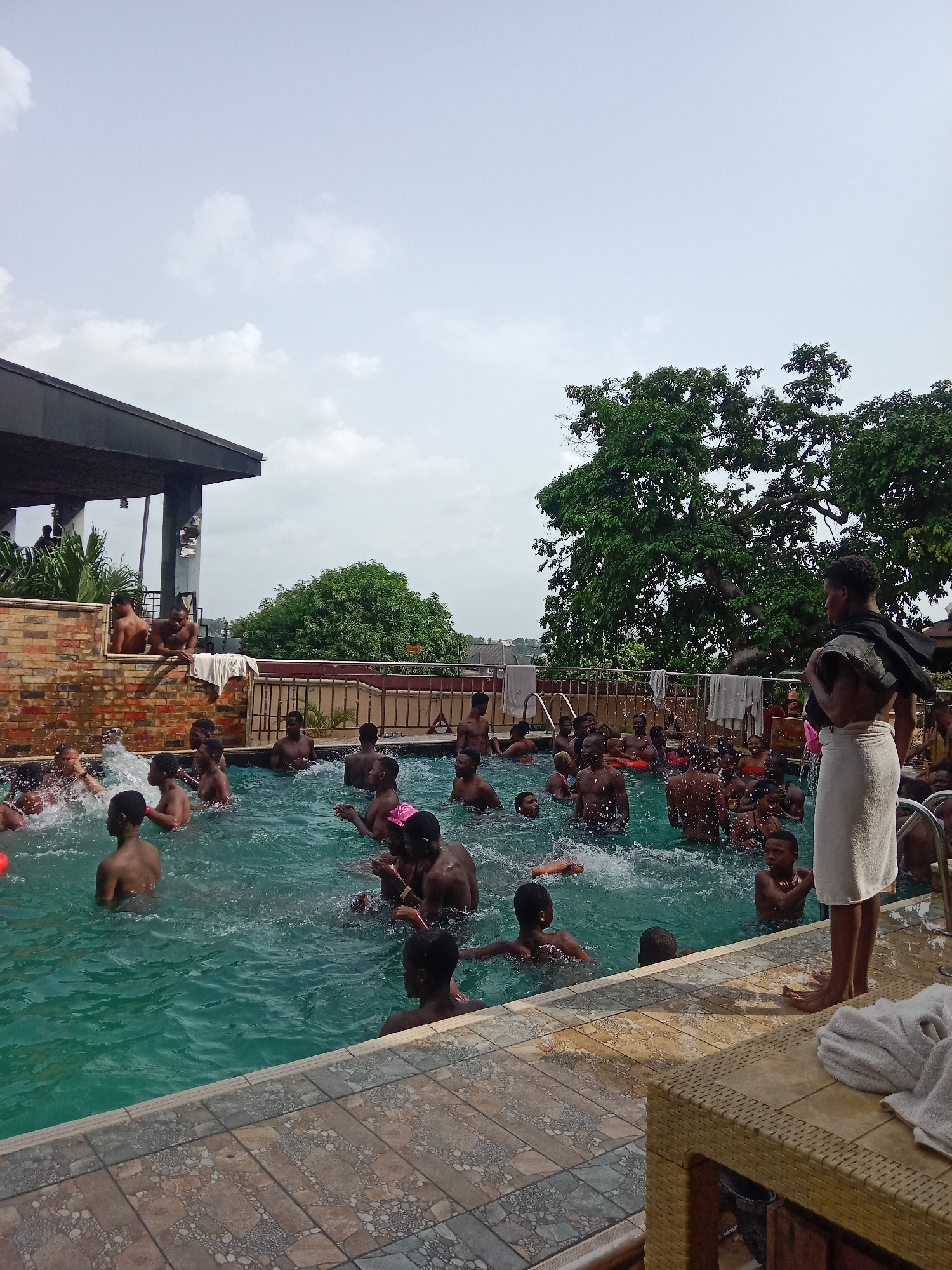 The image depicts a crowded outdoor swimming pool scene. The location is likely Onitsha, Nigeria, based on the subjects' presumed ethnicity and context. The pool is filled with numerous individuals, mostly young men and boys, who are swimming, splashing, and engaging in playful activities. One individual stands poolside wrapped in a white towel. A covered structure is seen behind the pool with additional people. Trees and an open sky are visible in the background. The pool's edge is tiled and there is patio furniture, including what appears to be a wicker table, is visible.