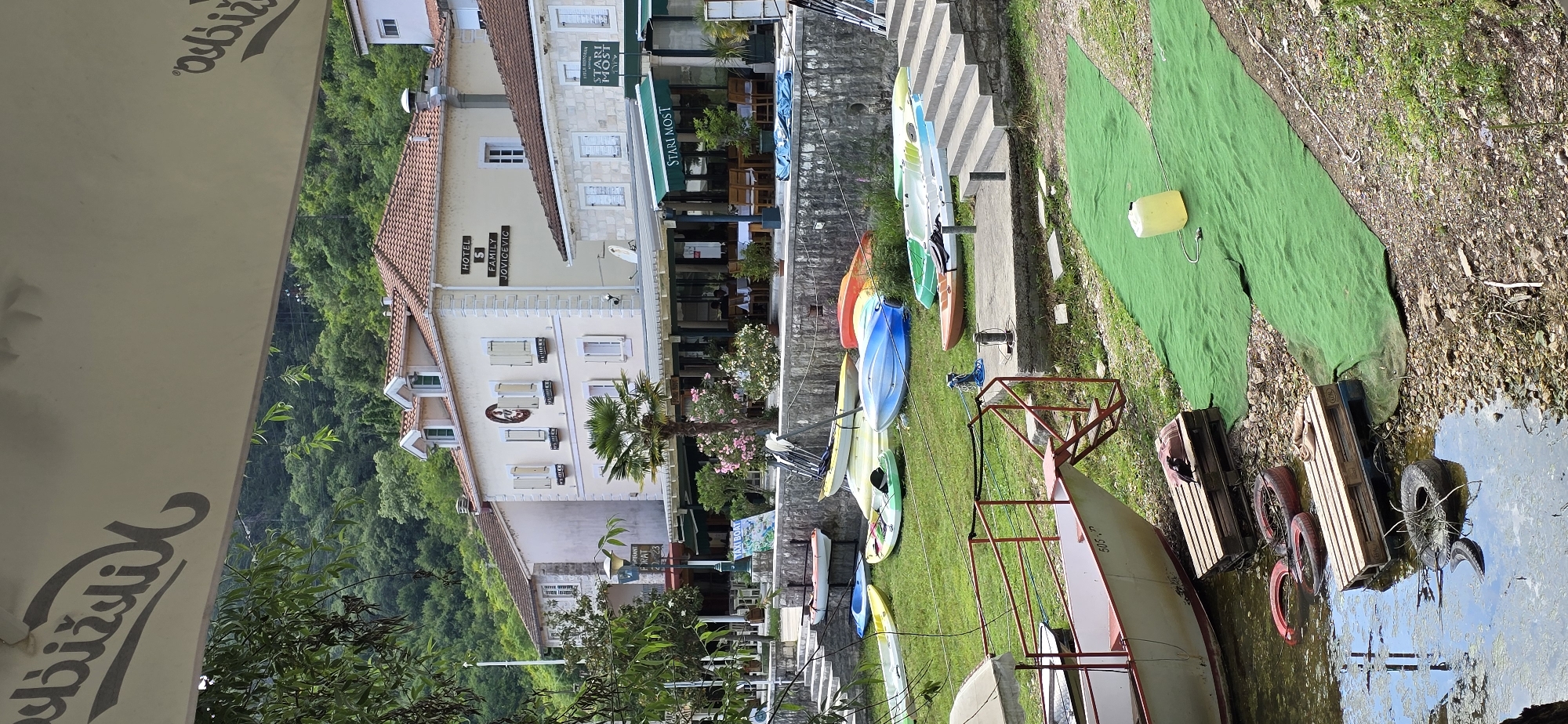 The image depicts a waterfront scene in Rijeka Crnojevića, Montenegro. In the foreground, a white and red boat is partially submerged in the water, with wooden pallets and tires scattered along the shoreline. Adjacent to the water, a vibrant green carpet covers an area of grass, with a yellow plastic container placed on it. Further back, a collection of colorful kayaks and canoes are neatly arranged on the lawn, suggesting a rental or recreational area.
Behind the kayaks, a stone wall separates the grassy area from a paved walkway leading up a series of concrete steps. Beyond the steps, a row of buildings lines the waterfront. One building, a hotel, is prominently featured with a sign reading "HOTEL S FAMILY JOVICEVIC." Another building appears to be a restaurant with outdoor seating. The architecture is consistent with traditional coastal European buildings, featuring tiled roofs and light-colored facades.
Overhead, a large white awning partially obscures the top of the image, with text visible in a stylized script. The background is dominated by lush green trees and hills, indicating a mountainous or heavily forested environment surrounding the town. The overall impression is one of a peaceful, picturesque location, likely a tourist destination with amenities for water activities and dining. No people are clearly visible in the image, but the presence of outdoor seating at the restaurant implies potential patrons. The scene is bathed in natural daylight, suggesting it was taken during the day.