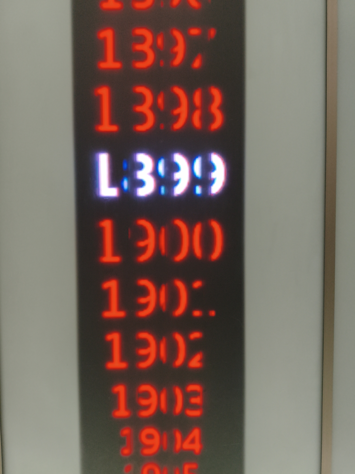 The image displays a digital LED sign showing a list of numbers, predominantly in red, with one line in white and blue. The numbers appear to be sequential, starting from 13:97 and continuing with 13:98, L:3:99, 19:00, 19:01, 19:02, 19:03, and 19:04. The sign is mounted vertically on a dark surface, which is casting a shadow on the right side. The background is a light-colored wall. The context of this sign's location in Ingolstadt, Germany, is not provided by the image itself.