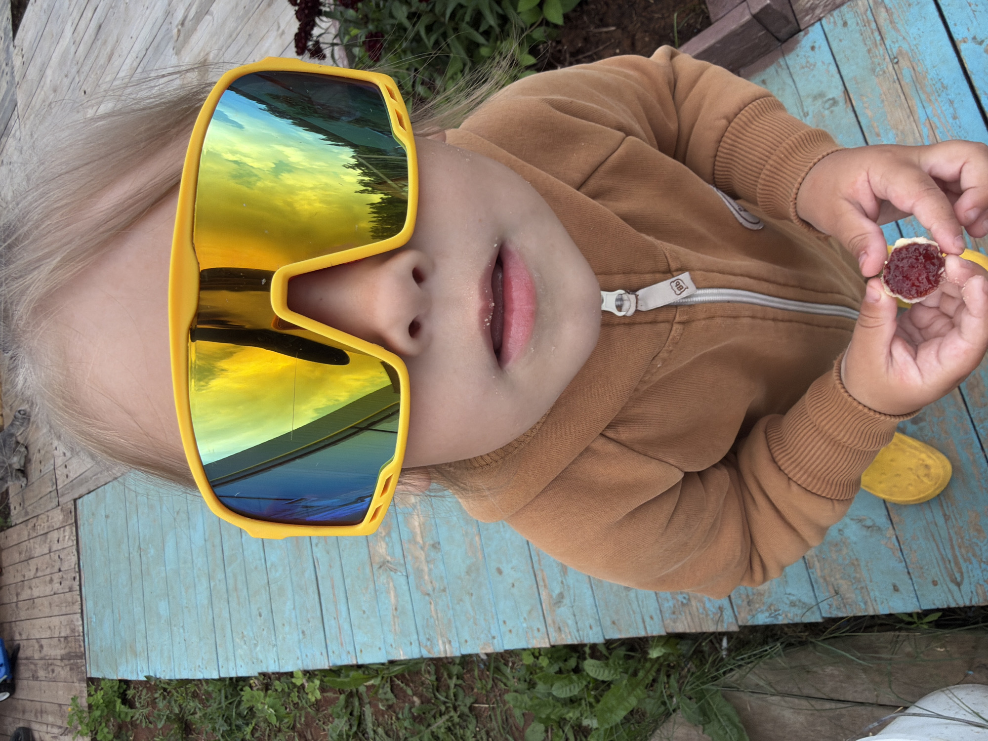 A young child wearing large yellow sunglasses and a brown hooded jacket stands on a blue wooden walkway. The child is holding a small round object with red jam on it in their hands. The walkway is surrounded by green grass and what appears to be a wooden deck or patio in the background. The image is taken from a high angle, looking down at the child.