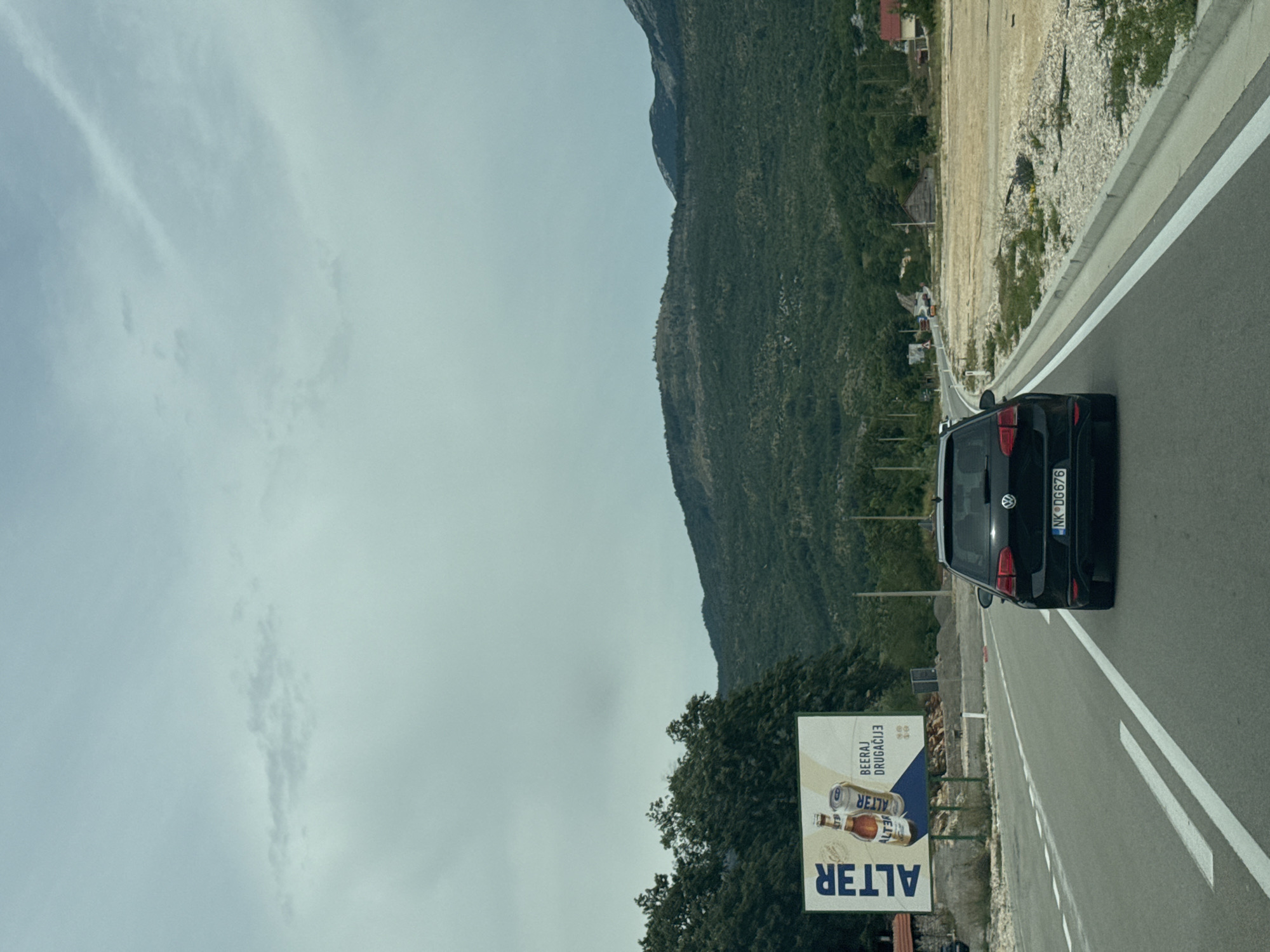 The image captures a view from inside a vehicle driving on a two-lane road in Jasenovo Polje, Montenegro. A black Volkswagen Golf, license plate NK DG676, is in the foreground, traveling in the same direction. To the left of the road, a large billboard displays an advertisement for "ALTER" beer, featuring two bottles and cans. The billboard is partially obscured by trees. The road curves gently to the right, leading towards a background of densely forested hills and mountains under a cloudy sky. On the right side of the road, there is an undeveloped area with dry grass and gravel, leading to scattered buildings. The scene suggests a rural or semi-rural setting with moderate vegetation and a vast, overcast sky.