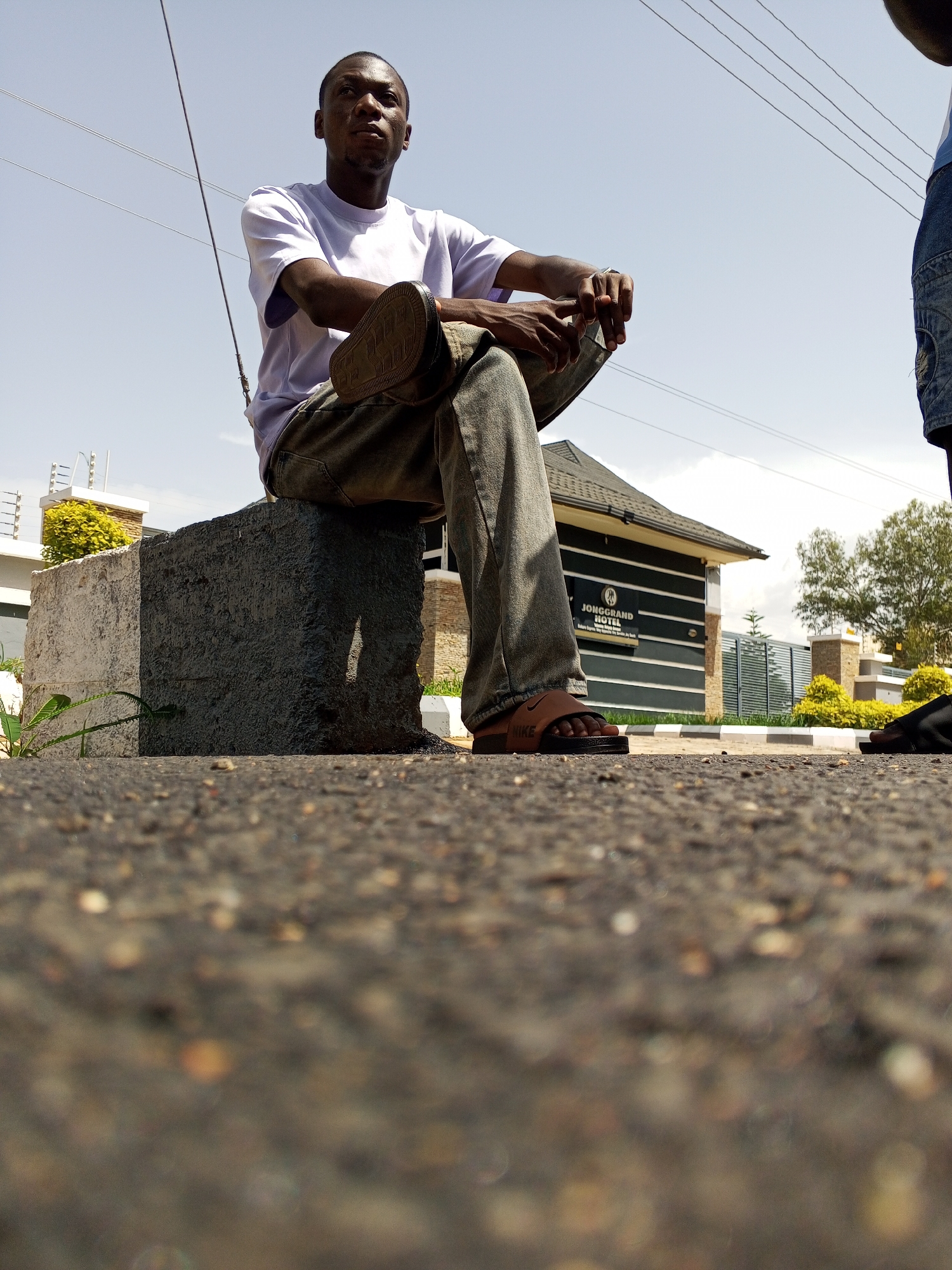 A low-angle shot captures a young man seated on a concrete block in Bukuru, Nigeria. He wears a light purple t-shirt, faded grey jeans, and brown slide sandals. His right leg is crossed over his left, with his right foot resting on the block. His hands are clasped in his lap. The background features a building with "JONGGRAND HOTEL" signage, manicured hedges, and trees under a bright, hazy sky with visible power lines. The foreground consists of asphalt pavement. Another person's legs and feet are visible to the right, partially cut off by the frame.