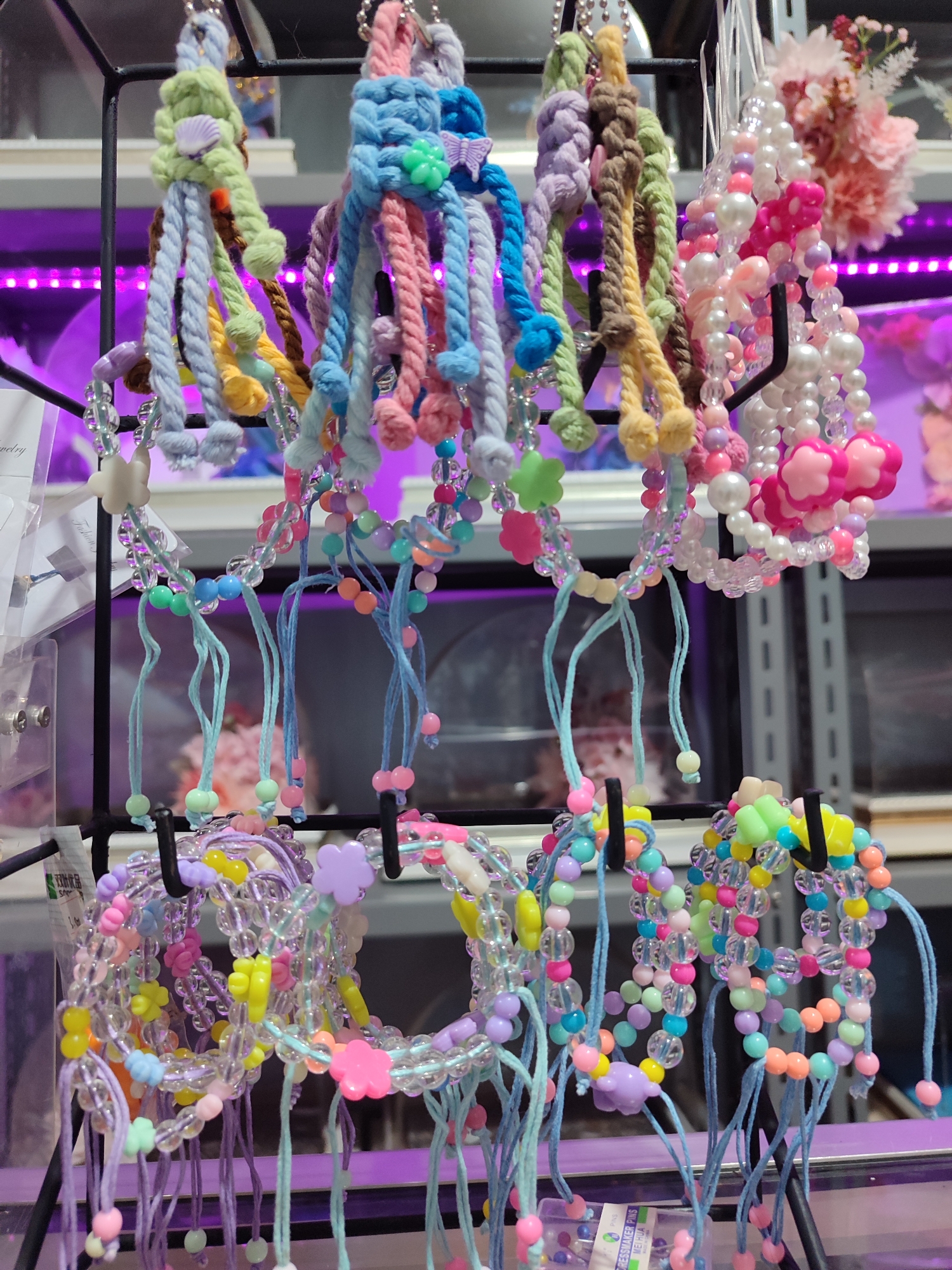 A vertical, medium shot captures a display of colorful, handmade beaded bracelets and knitted keychains, likely for sale in a market or shop in Gantungan, Indonesia. The items are suspended from a black metal rack, illuminated by bright, artificial purple lighting.
The foreground is dominated by rows of bracelets, made from clear, iridescent beads interspersed with brightly colored plastic beads in shades of pink, blue, green, yellow, and orange. Many of these bracelets feature small, flower-shaped charms in pastel colors. Dangling from some of the bracelets are thin, light blue strings with single beads at the ends.
Above the bracelets, a row of knitted keychains hangs. These keychains are made of thick, braided yarn in various pastel colors, including light green, blue, purple, and pink. Some of these keychains are adorned with small charms such as seashells, butterflies, and floral shapes.
In the background, shelves are visible, also lit with the purple light, suggesting a retail environment. A cluster of artificial pink flowers is visible on the upper right side of the frame, adding a decorative element. The overall impression is one of a vibrant and cheerful display of handcrafted accessories.