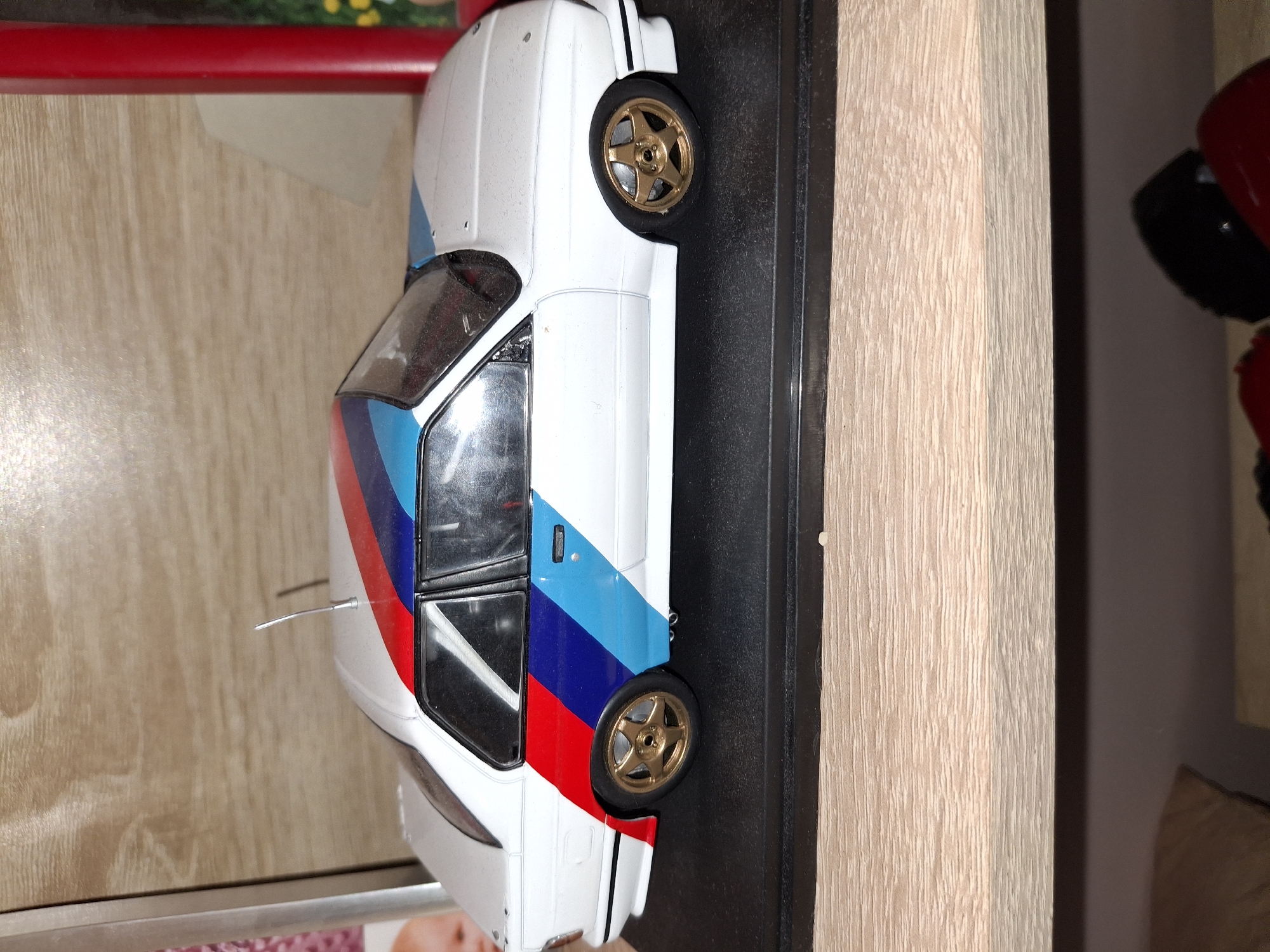 A model car, white with blue, red, and darker blue racing stripes, is displayed on a black base. The car has gold wheels and tinted windows. A thin, silver antenna extends from the rear of the car. The car is positioned on a shelf or display surface with a light-colored wood-grain texture. In the background, a red bar is visible above the shelf, and to the right, a blurred object with a red top and black bottom, possibly another collectible, is partially seen. The lighting suggests an indoor setting. The image appears to be taken from a slightly elevated angle looking down at the model car.