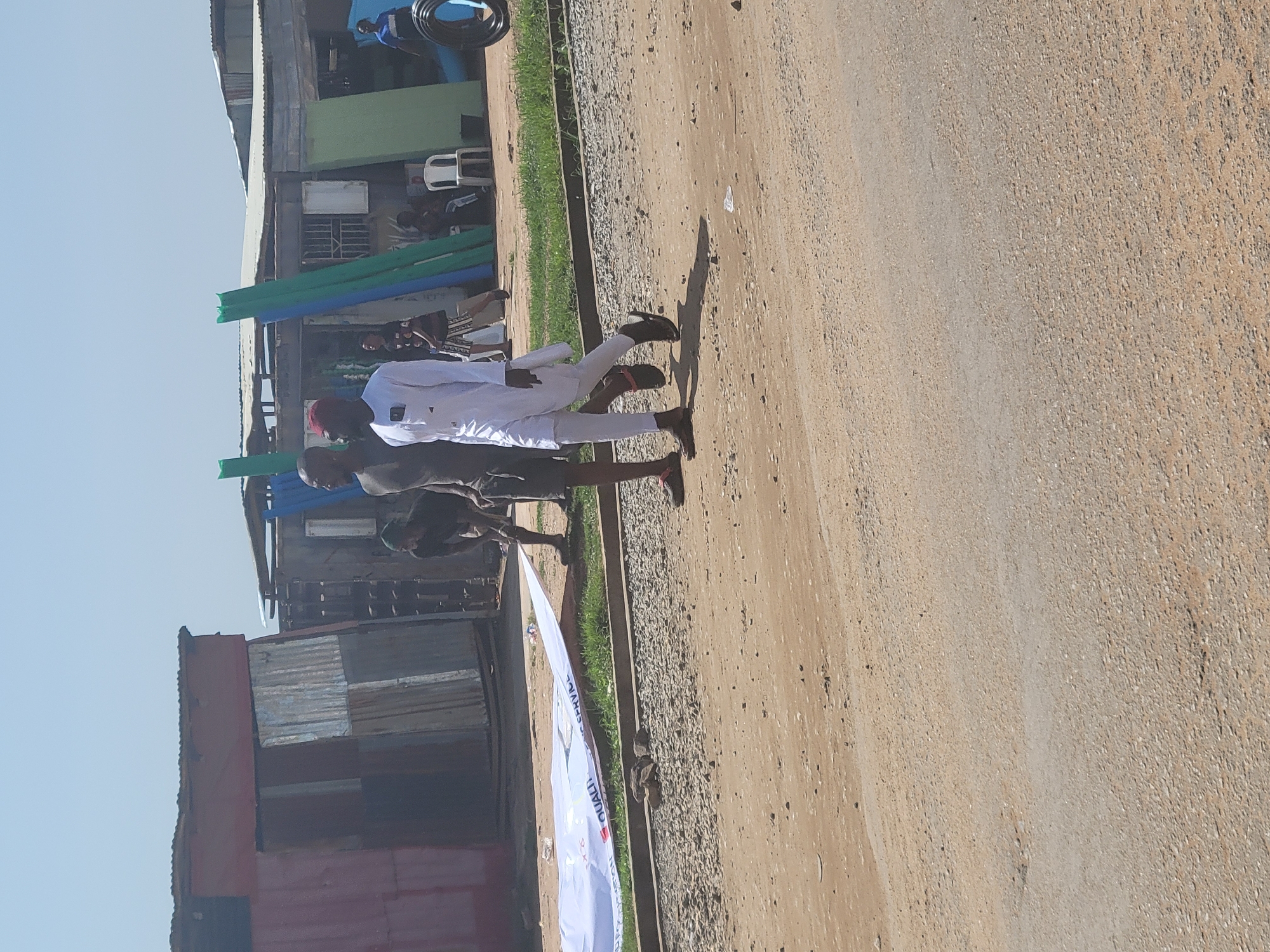 In Kwamba, Nigeria, two men walk on a dirt road adjacent to a row of makeshift structures constructed from corrugated metal. The man on the left wears a dark t-shirt and shorts, while the man on the right is dressed in a white traditional garment and a red cap. They are walking towards the right side of the frame. In the background, a large white banner with text is partially visible, lying on the ground. The structures in the background appear to be shops or informal dwellings, with various materials like metal sheets and tarps used for their construction. The sky is clear and bright, indicating a sunny day.