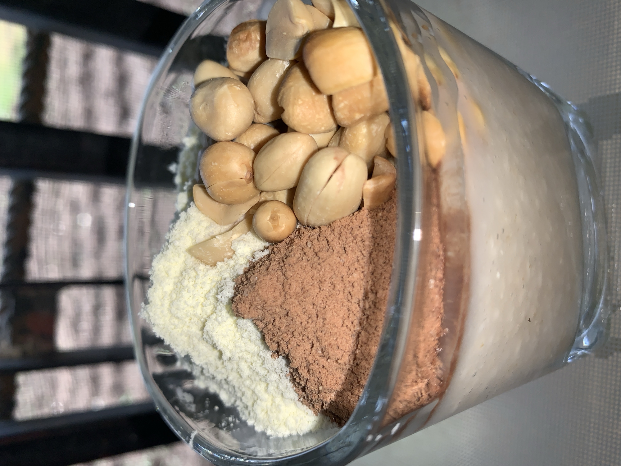 The image shows a glass filled with multiple layers of food items. At the top, there is a heap of shelled peanuts. Below the peanuts, there's a layer of white powder, next to a portion of brown powder. The remaining space in the glass is filled with a beige, liquid-like substance. The glass appears to be indoors, with a blurred background showing a window or similar structure. There is no information available regarding the location of the image in the city of Rafin Par, Nigeria.