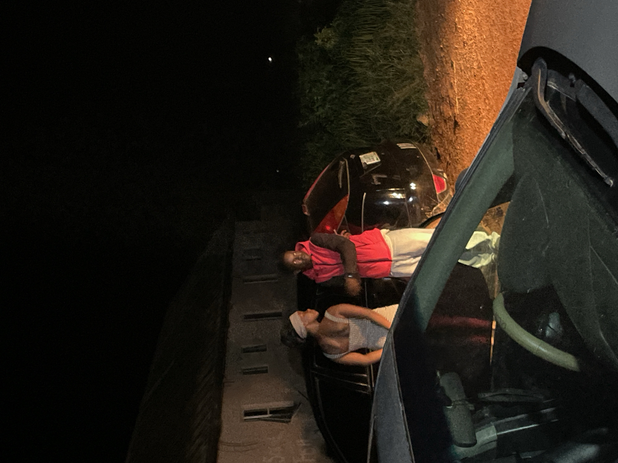 In Wumat, Nigeria, at night, two individuals, a woman and a man, stand near parked vehicles. The woman, facing away from the camera and to the left, wears a striped dress and a headscarf. The man, facing the camera and to the right, is dressed in a red vest over a dark shirt and white pants. They are positioned between a building with multiple windows and a dark-colored car. The foreground is occupied by the hood and windshield of another vehicle. The environment appears to be an outdoor, possibly rural setting, with vegetation visible in the background and a dirt ground surface. The scene is illuminated by artificial light sources, casting shadows and creating a dramatic atmosphere.
