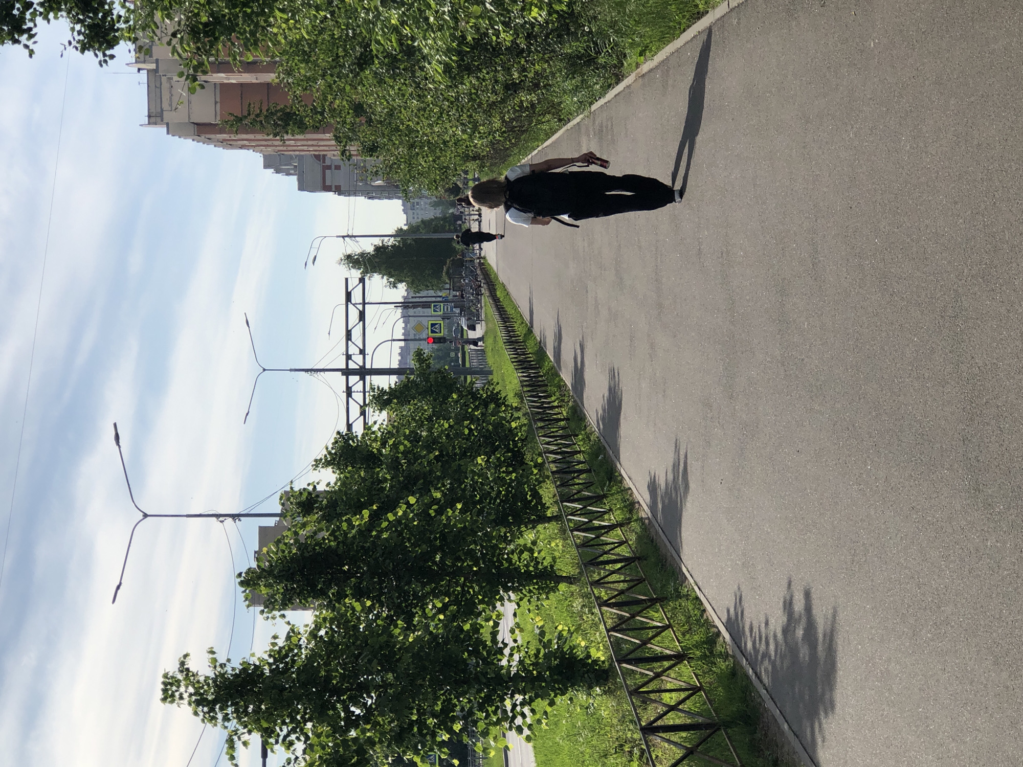This detailed description pertains to an image captured on a sunny day, likely during late spring or summer, in an urban area of Saint Petersburg, Russia. The scene depicts a straight asphalt pedestrian and cycling path receding into the distance, flanked by greenery and urban infrastructure.
On the left side of the path, a low, dark metal fence with a criss-cross pattern separates the pathway from a narrow strip of green grass. Beyond this grass strip, a uniform row of mature deciduous trees with lush green foliage forms a visual barrier, indicating separation from a wider multi-lane asphalt road. Sunlight filters through these trees, casting dappled light and distinct shadows on the grass and the fence. Further down the street on the left, an intersection is visible with a red traffic light for vehicles and a distinct white triangular pedestrian crossing sign. Overhead, a network of wires and tall, dual-arm streetlights traverse the scene.
On the right side of the path, a dense hedge of green bushes and smaller trees runs parallel to the path, providing a natural boundary. Beyond this hedge, in the mid-ground and background, stand multi-story residential buildings, characteristic of Soviet-era construction common in Saint Petersburg. These buildings feature facades with alternating sections of reddish-orange brickwork or panels and lighter-colored (possibly cream or white) sections.
In the immediate foreground, a person with light-colored hair, wearing a white t-shirt, dark trousers, and a black backpack, walks away from the camera along the path. This individual holds a red and silver can in their right hand. Their elongated shadow is cast forward on the asphalt path, suggesting the sun is relatively low in the sky, either morning or late afternoon. Further along the path, another person is visible riding a kick scooter, also moving away from the camera. Several other indistinct figures appear in the far distance on the path, suggesting a moderate level of pedestrian activity.
The sky above is light blue with scattered, wispy white clouds, indicating fair weather. The overall environment appears clean and well-maintained, typical of a residential or park-adjacent urban thoroughfare in Saint Petersburg.