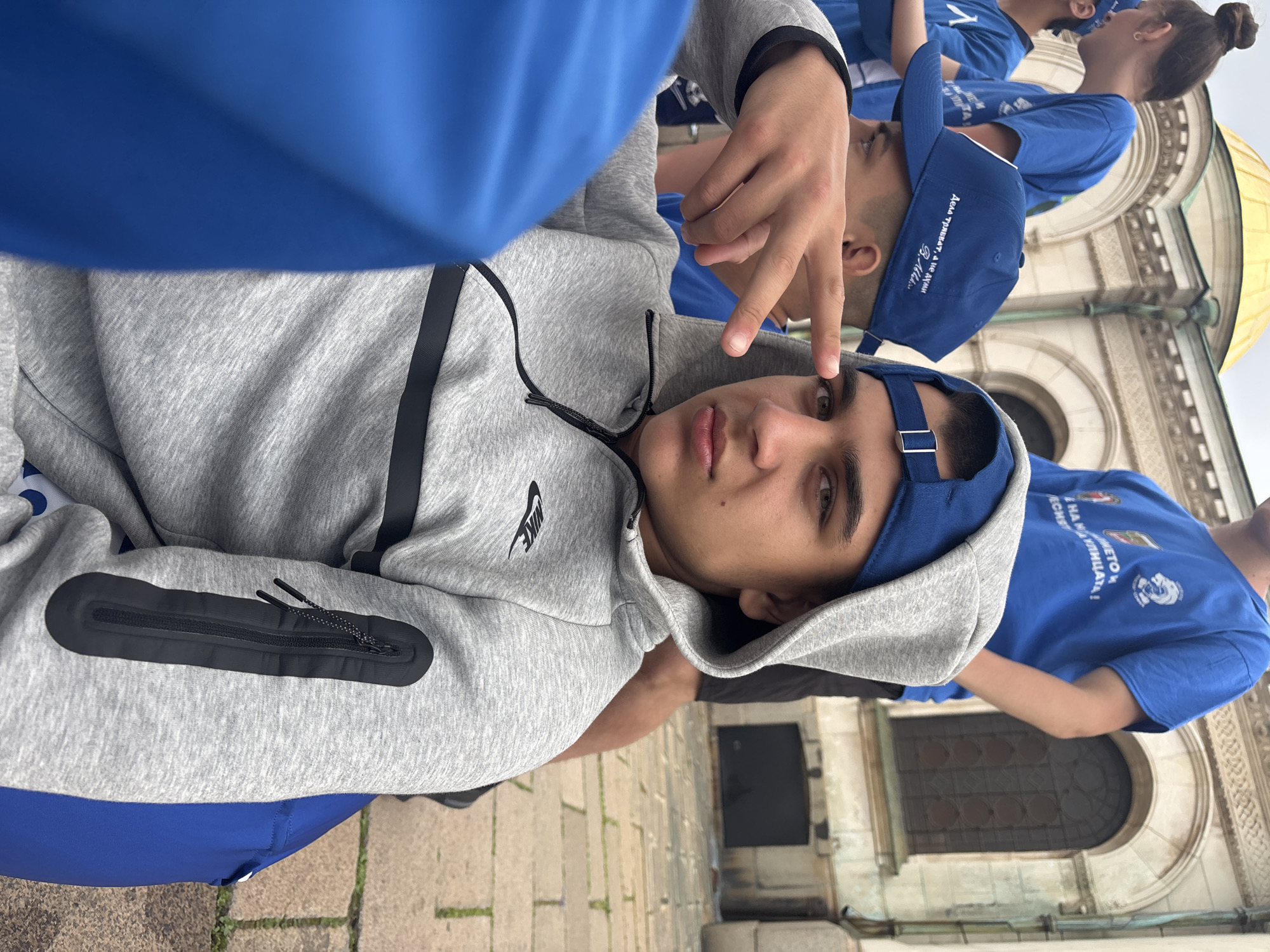 A low-angle shot shows a young man in a gray Nike hoodie and a blue cap, making a hand gesture. Behind him are other young people wearing blue t-shirts and caps. The location is Sofia, Bulgaria, as evidenced by the Cyrillic script on the caps and t-shirts and the architectural details of the building in the background, which resembles Alexander Nevsky Cathedral.