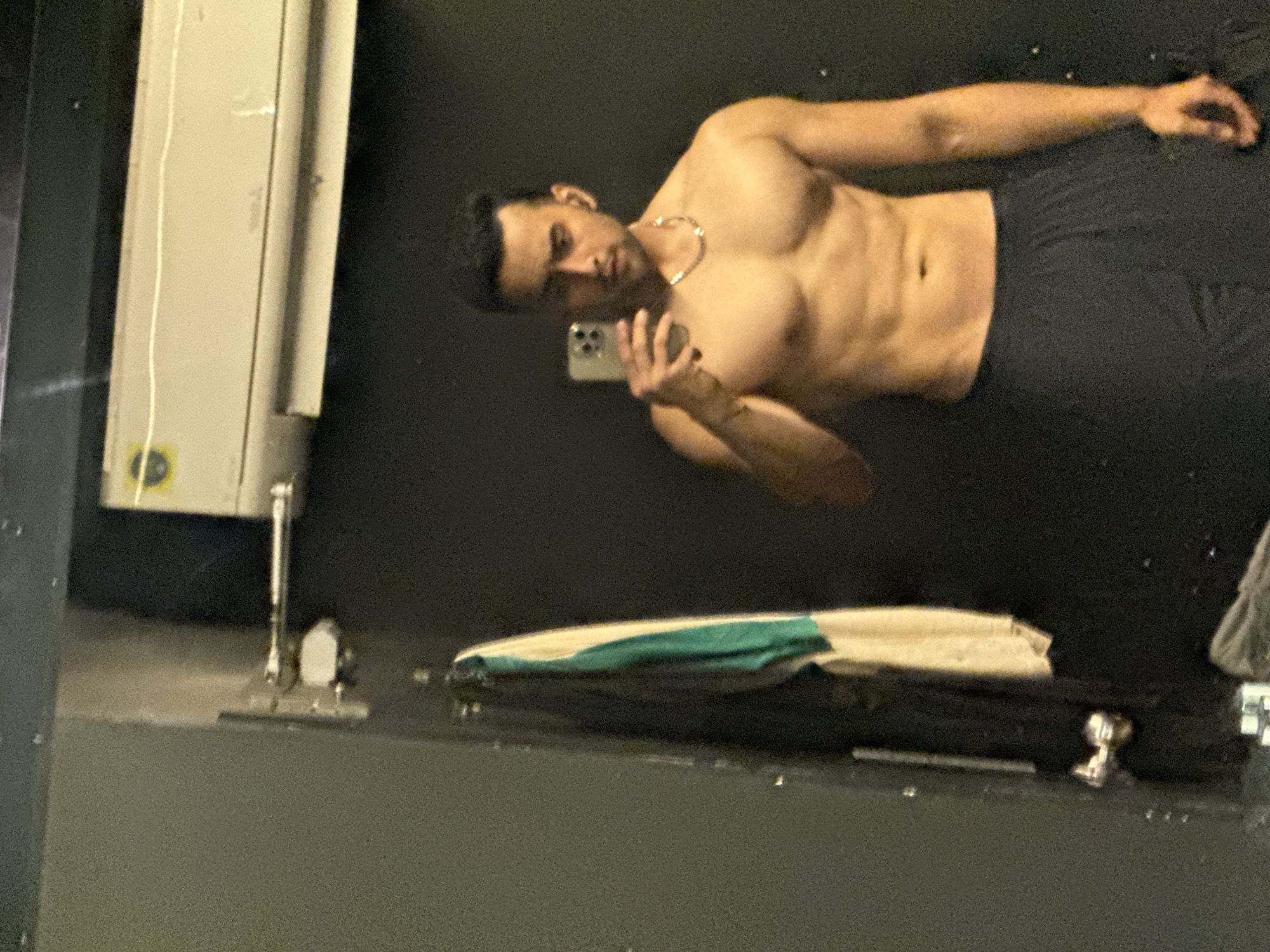 A shirtless man with dark hair is taking a selfie in what appears to be a gym. He's wearing black shorts and a chain necklace. He's holding a phone in his left hand and looking at the camera. In the background is a gray wall and a piece of gym equipment. There is a towel on the equipment. There is nothing in the image to indicate it is Kolkata, India.