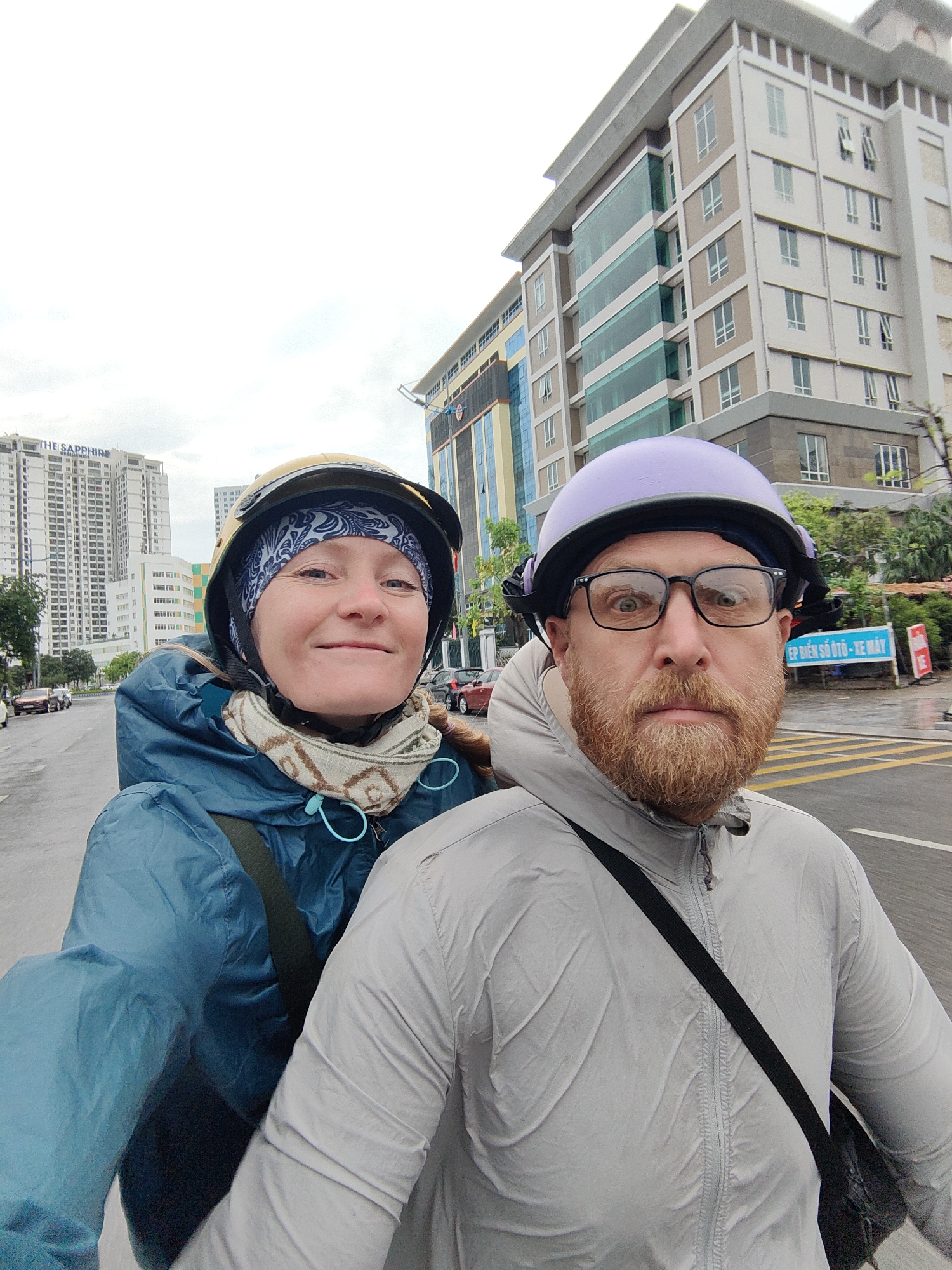 A woman and a man are riding a motorbike on a city street, as observed in Hong Gai Ward, Vietnam. The woman, positioned behind the man, is wearing a dark blue head wrap under a golden helmet and a blue raincoat. She has a scarf around her neck. The man, in front, has a red beard, eyeglasses, and a purple helmet. He is wearing a grey jacket with a black strap across his chest. Both individuals appear to be taking a selfie or having someone take their picture. The street shows signs of wet conditions, possibly from recent rain. Background elements include buildings, including "The Sapphire" high-rise apartment complex. A Vietnamese sign is visible near the side of the street.