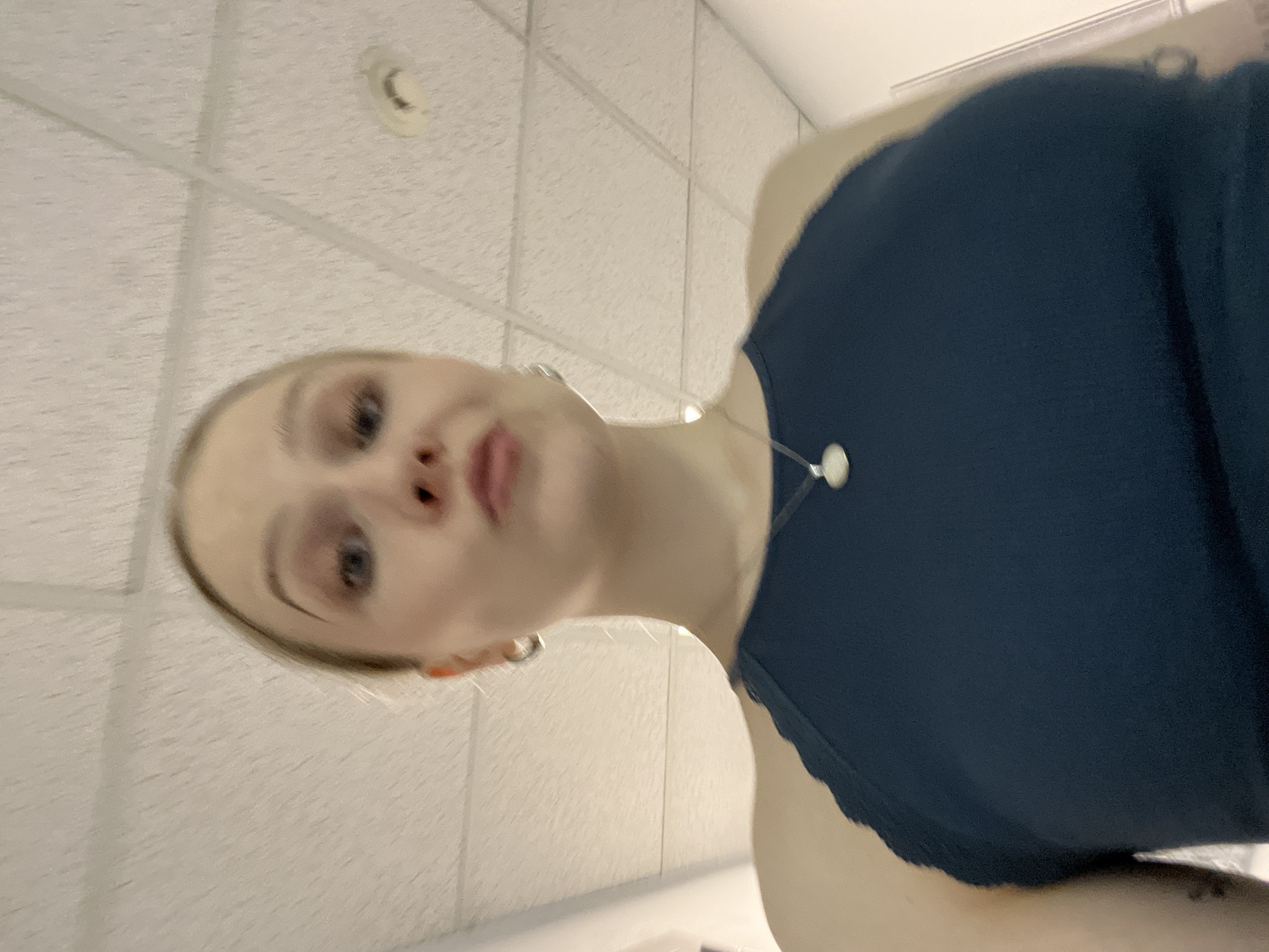 A woman with blonde hair, fair skin, and blue eyes is angled upwards towards the camera. She is wearing a dark blue top with lace trim and a silver necklace. The background consists of a white tiled ceiling with a circular fixture. The location is not discernible from the provided image.