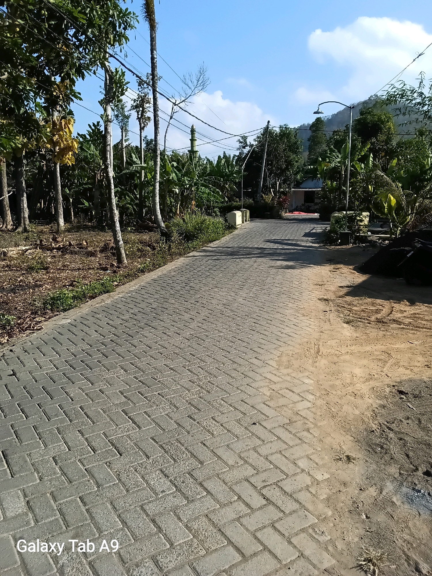 The image shows a paved pathway in a rural or semi-rural area. The pathway is made of grey rectangular pavers laid in a herringbone pattern. To the left of the path, there is an area with dry leaves, dirt, and trees with a few banana trees visible. To the right of the path, there is a sandy area with tire tracks and a pile of dark material, possibly soil or construction debris. In the background, there are more trees and vegetation, a few small buildings, and a street light. A hill with vegetation is visible in the distance under a bright blue sky with some white clouds. The watermark "Galaxy Tab A9" is visible in the bottom left corner. The scene appears to be a street or access road within a residential or agricultural area. Based on the context of Trenggalek, Indonesia, this could be a village road or a pathway leading to a house or farm.