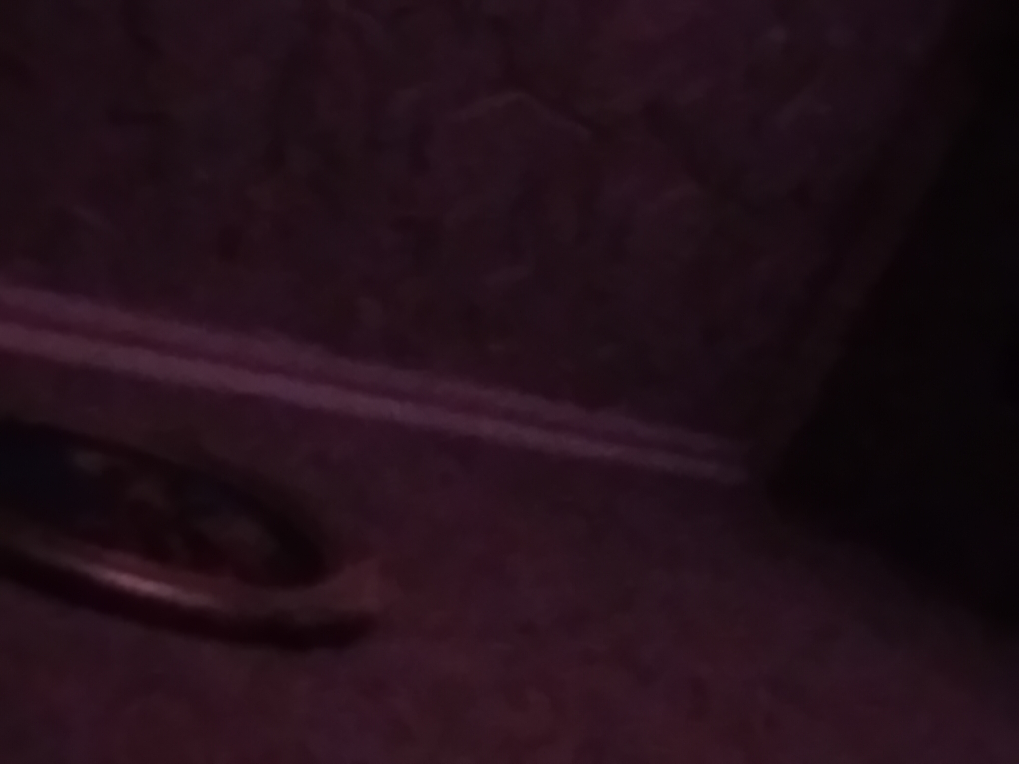 A close-up, blurry, and dimly lit image shows a portion of a wall with a textured pattern, possibly hexagonal, in a dark purple hue. A vertical, lighter purple line runs down the right side of the image, suggesting a corner or a seam. In the upper left corner, a circular object with a metallic rim is visible, partially obscured and out of focus. It is impossible to determine the exact nature of this object or its surroundings with the given visual information. The image does not contain any discernible features related to people, actions, or interactions.