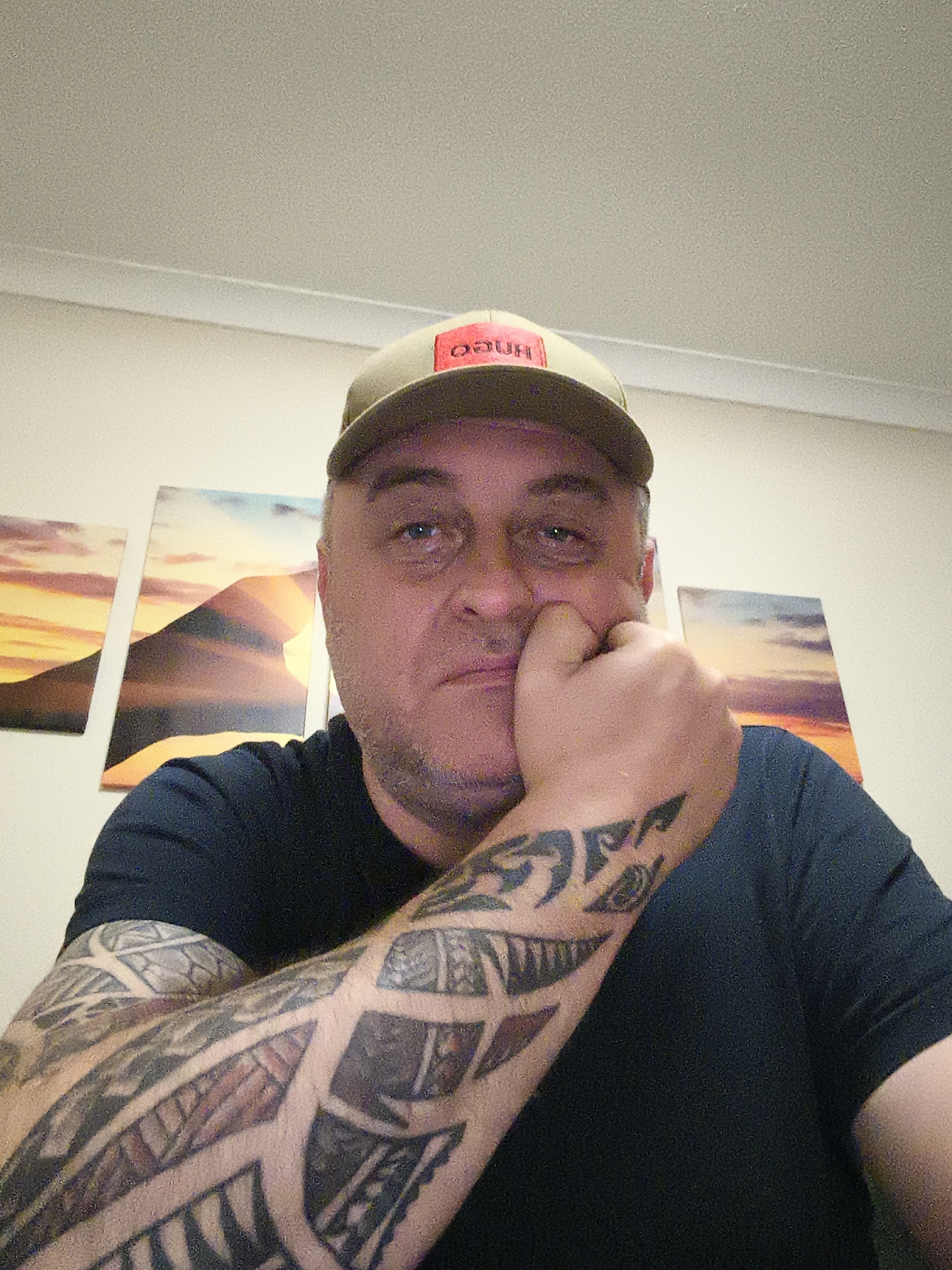 A man is depicted indoors, likely in a room with light-colored walls and a white ceiling. He is wearing a dark t-shirt and a light-colored baseball cap with a red label on the front. On this label, the word "HUGO" is visible, appearing reversed, possibly due to a front-facing camera's mirroring effect. He has extensive dark, tribal-style tattoos covering his right forearm and parts of his left arm. The man is posing for the photo, resting his left hand, clenched into a fist, against his cheek with a pensive or serious expression. His gaze is directed towards the camera. In the background, on the wall, there are three rectangular art pieces, positioned side-by-side, depicting a sunset or sunrise over mountainous landscapes. The image is taken from a low angle, looking up at the man.