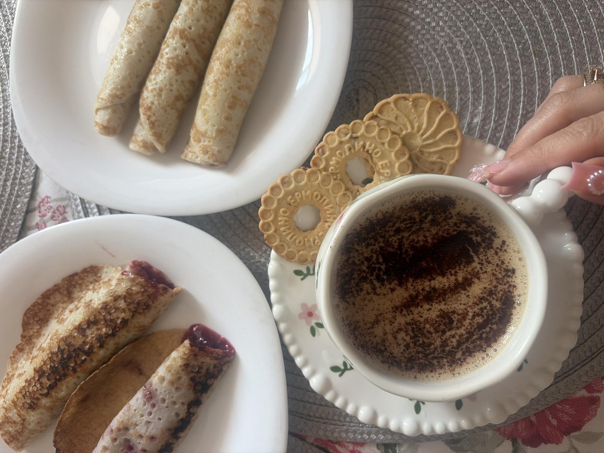 The image displays a meal setup on a table. A gray woven placemat rests atop a floral-patterned fabric with red and pink elements. Two white ceramic plates are visible. The left plate contains two crepes; one is rolled and exhibits a dark red, presumably fruit-based, filling visible at both ends and through its textured, slightly browned surface. The second crepe on this plate is partially rolled, also revealing a similar dark red filling. The right plate holds three uniformly rolled, light golden-brown crepes. In the foreground, a white ceramic teacup with a scalloped-edge saucer, decorated with pink flowers and green leaves, contains a beverage topped with foam and dark powder. Three round, light-colored biscuits with an embossed pattern are positioned next to the cup; one biscuit distinctly displays the Cyrillic text "ЕРМОЛИНО" (Yermolino), identifying a Russian food brand. A female hand, featuring pink, embellished artificial nails and a gold-colored ring on the ring finger, is shown holding the handle of the teacup. The presence of the "Yermolino" brand biscuits indicates the location is in Russia, consistent with the city of Saint Petersburg.