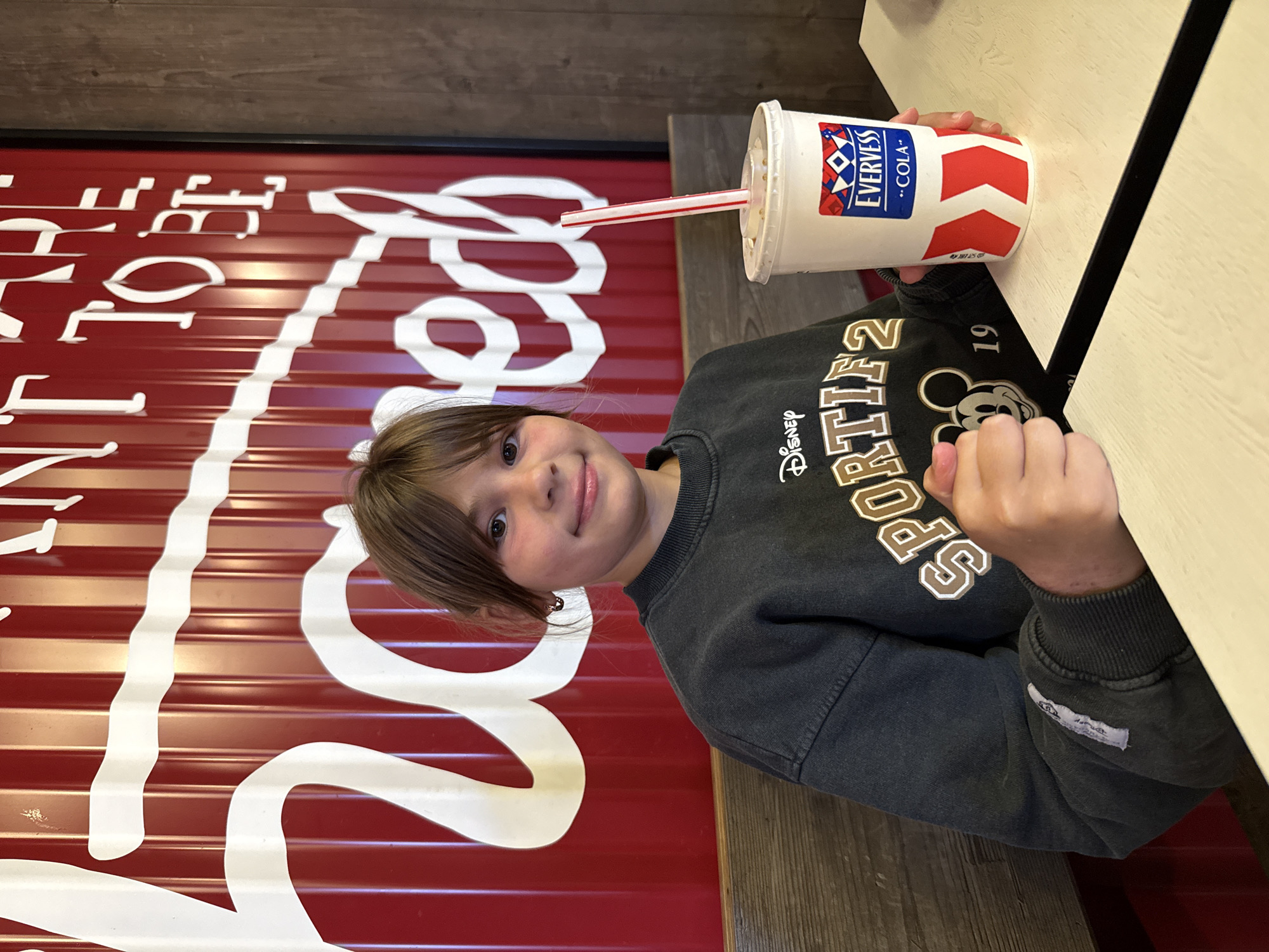 A young girl is seated at a table in what appears to be a fast-food restaurant, possibly in Ignatovo, Russia. The background features a prominent red corrugated metal wall with large white lettering that reads "WANT TO BE". The girl is wearing a dark gray sweatshirt with a Disney Mickey Mouse graphic and the word "SPORTIF" on it. She is holding a white cup with a red and white striped straw and a logo for "EVERVESS COLA". The table surface is light-colored with a visible seam. The lighting suggests an indoor setting.