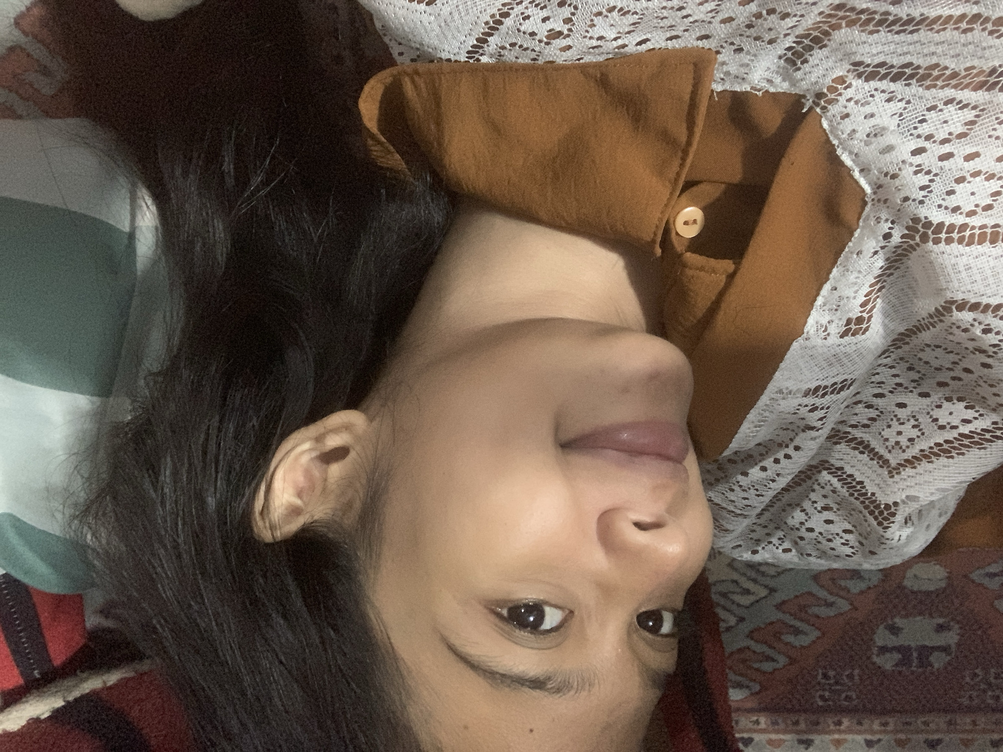 The image shows a close-up of a young woman's face as she lies down, possibly on a bed, in a room. Her head is tilted, and she is looking towards the viewer with a slight smile. Her dark hair is spread out around her. She is wearing a brown collared shirt with a white lace overlay. The background appears to be a patterned blanket or rug, with hints of red, brown, and cream colors. A green and white striped pillow is visible behind her head. There is no indication of the specific location within Sungai Meriam, Indonesia.