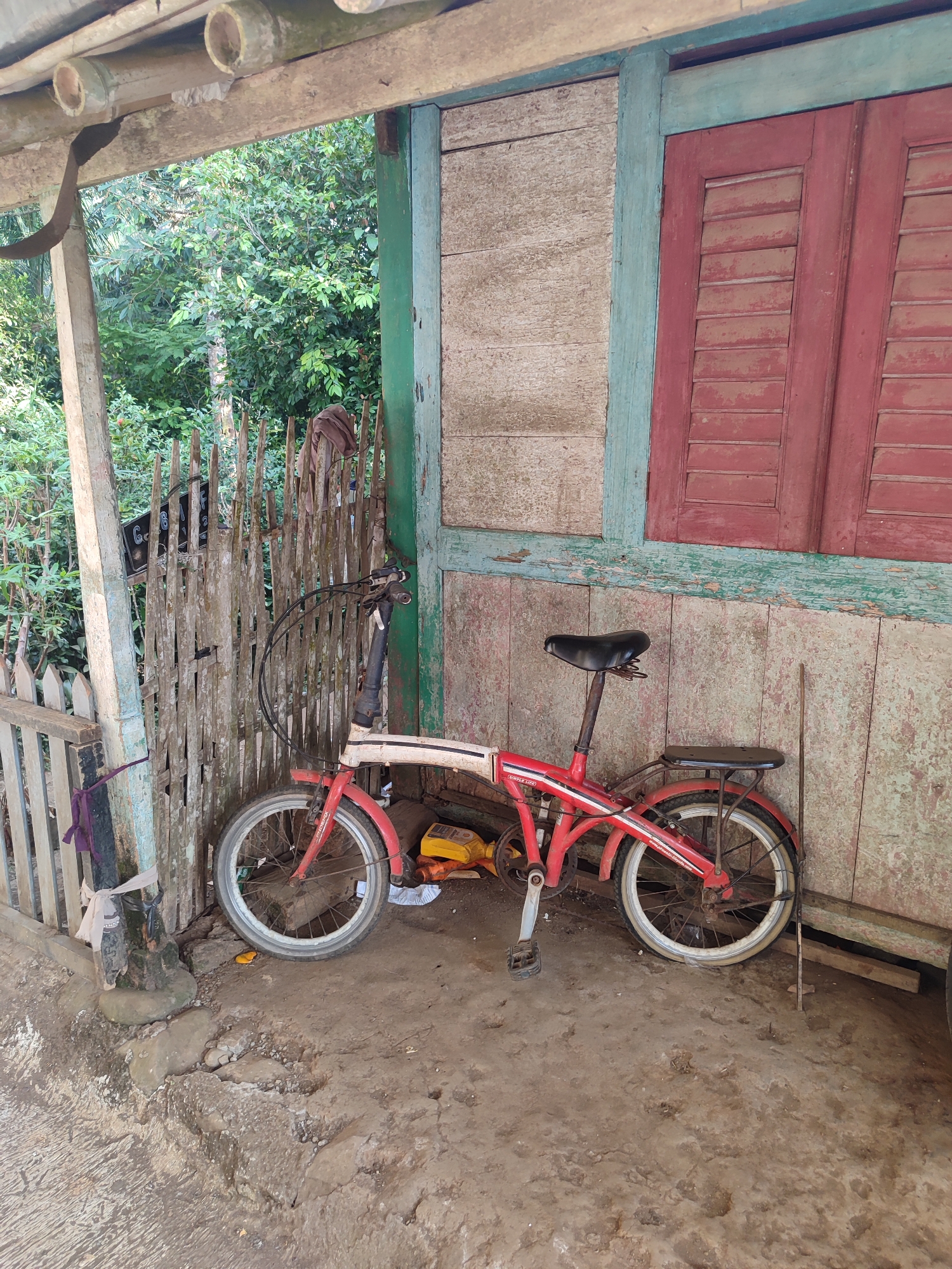 A weathered, red and white folding bicycle is parked against the side of a rustic wooden structure. The bicycle has a black seat and a rear rack. The building's walls are constructed of roughly textured wood planks, some painted in faded colors of light beige, teal, and red. A red shuttered window is visible on the right side of the building. In the background, a wooden fence stands partially obscured by lush green foliage, suggesting an outdoor setting in Gantungan, Indonesia. The ground is a mixture of dirt and concrete with some debris. There are no people visible in the image.